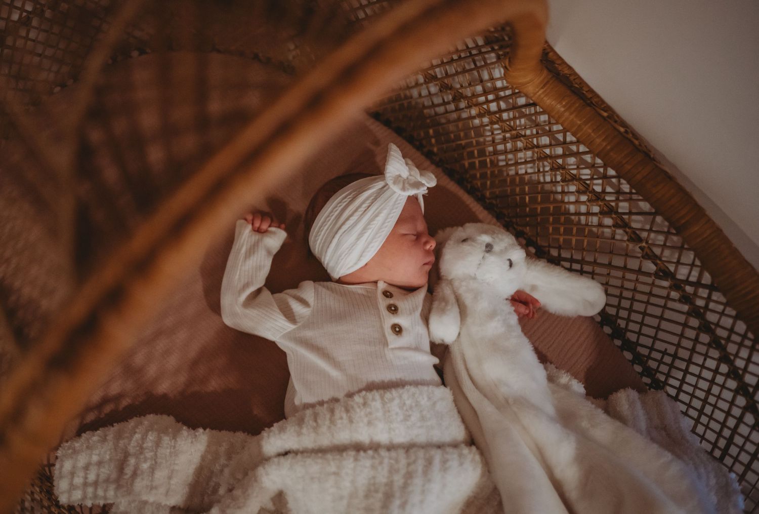 Sleeping newborn baby in white outfit and headband bow lying in a wicker bassinet with stuffed toy.