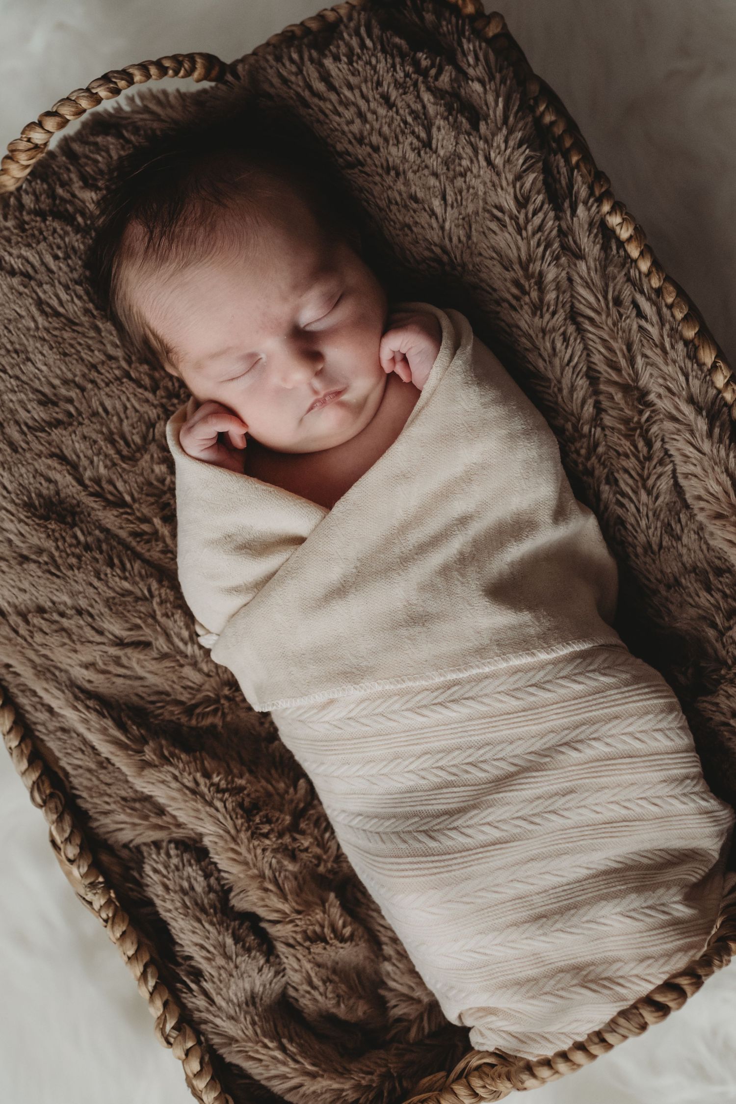 A swaddled newborn sleeps peacefully in a woven basket lined with a soft brown blanket.
