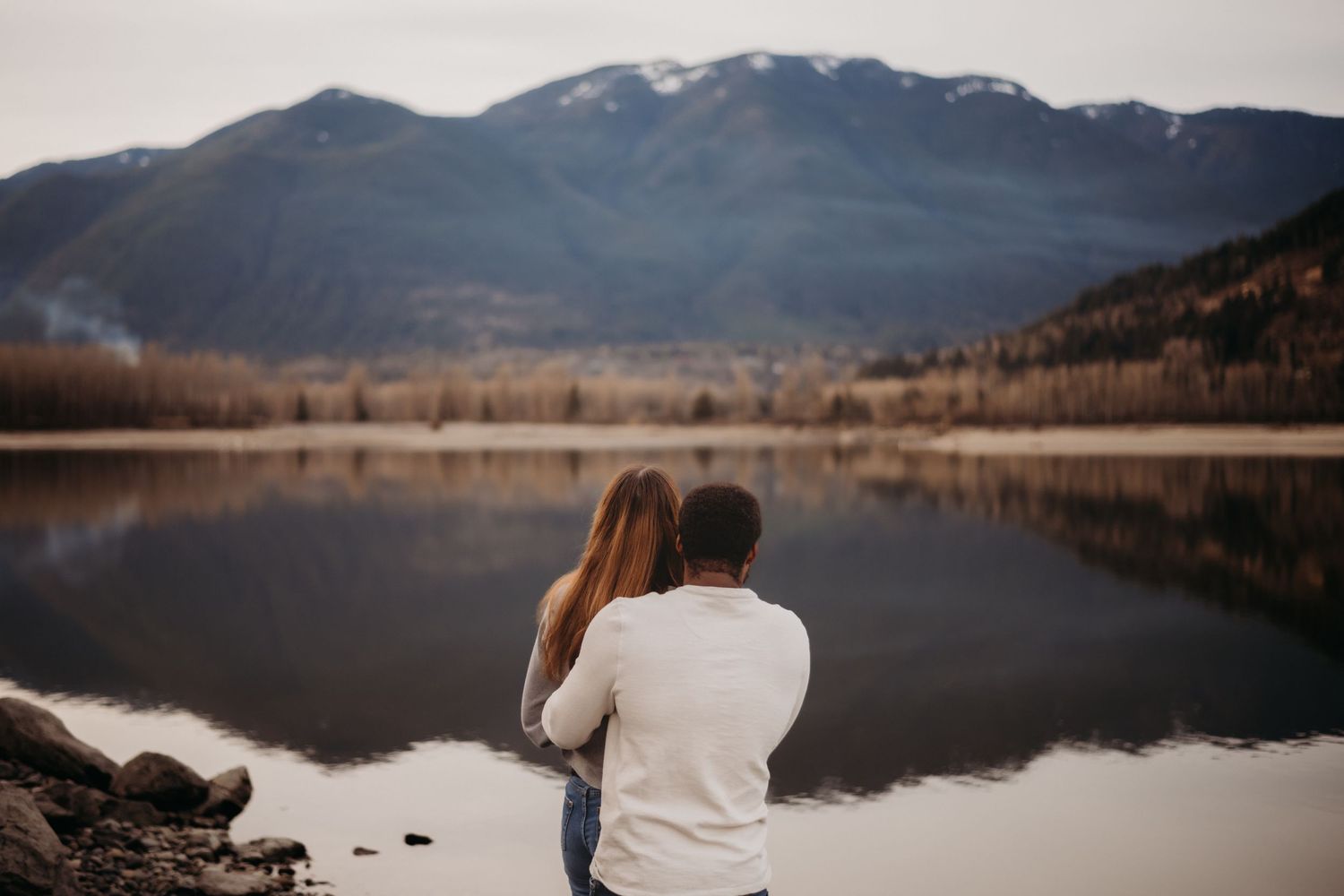 Interracial couple sitting together by a calm mountain lake in an intimate embrace.