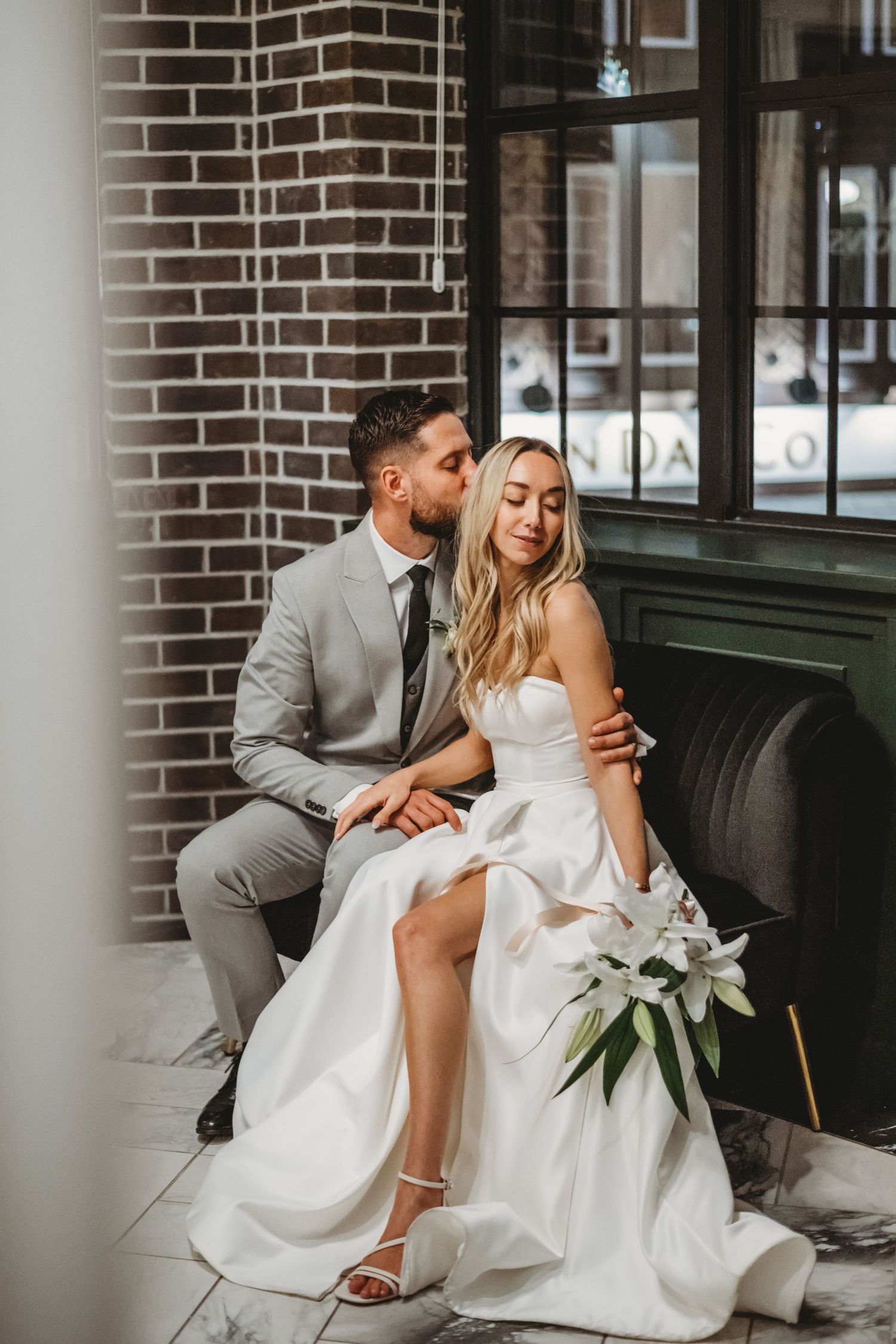 Groom gently kisses bride on cheek as she sits gracefully in a modern wedding venue.
