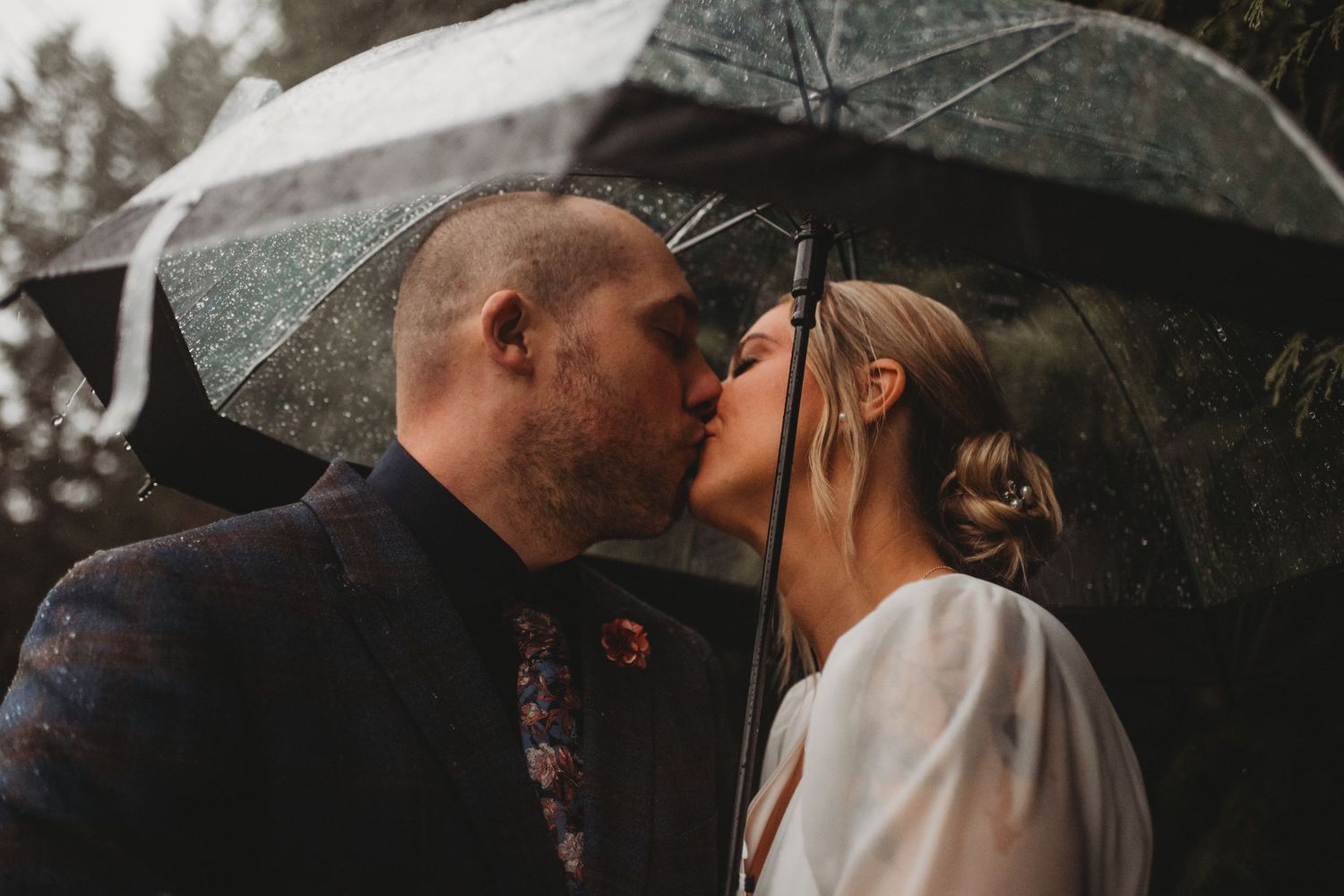 Bride and groom kissing under a clear umbrella in the rain during their outdoor wedding.