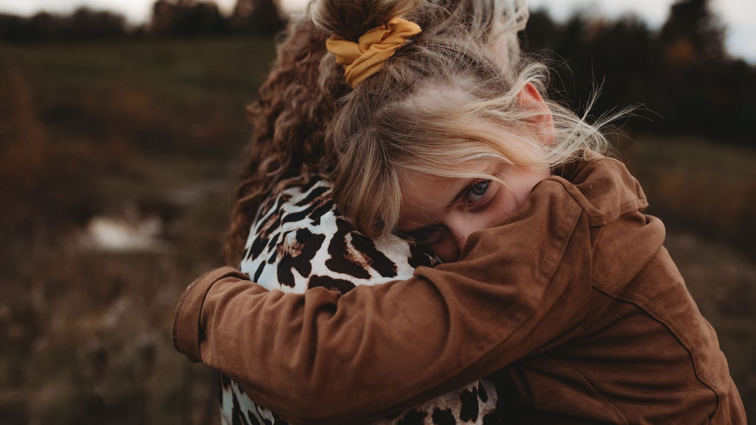 Young girl with yellow bow hugging woman in leopard print during outdoor fall photoshoot.