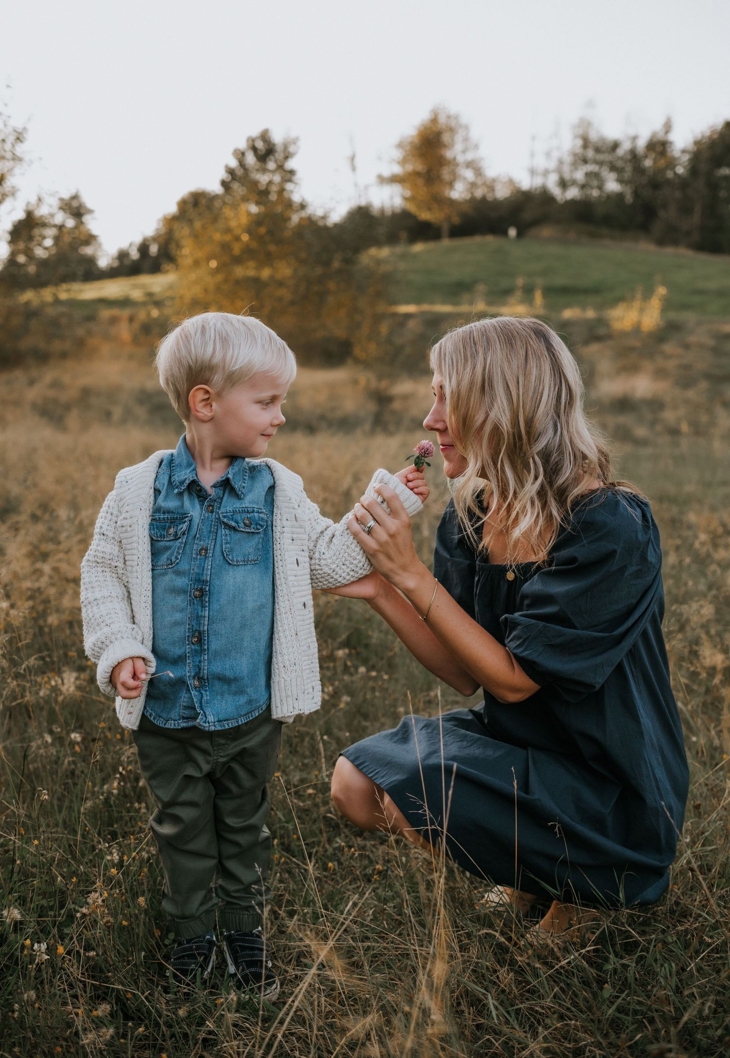 Blonde mother kneeling in field smelling flower offered by young son at golden hour.