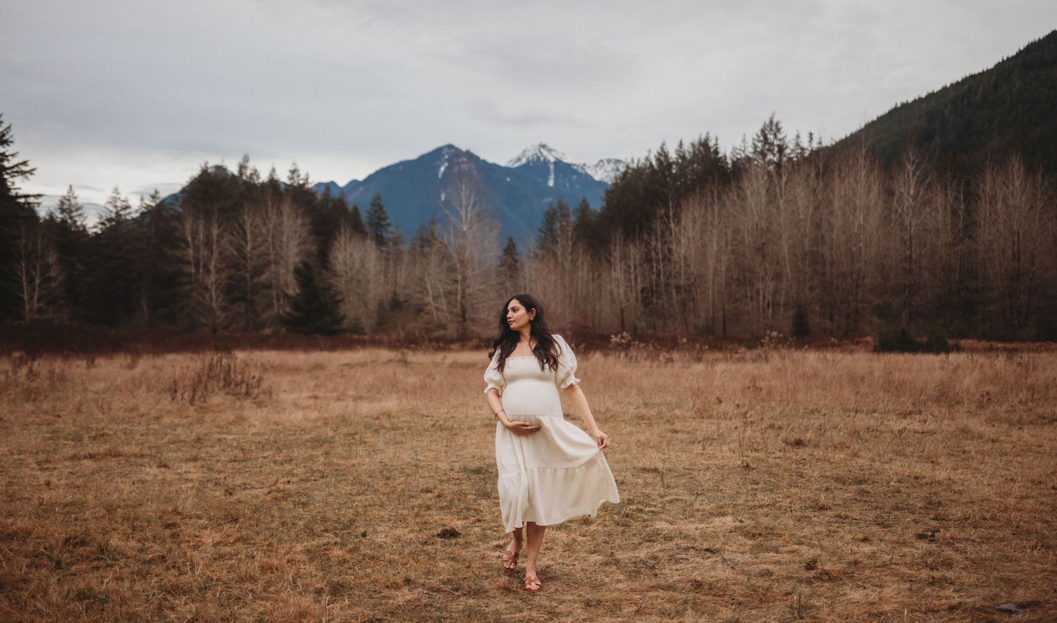 Expectant mother in flowing white dress twirls in autumn field with mountain scenery behind her.