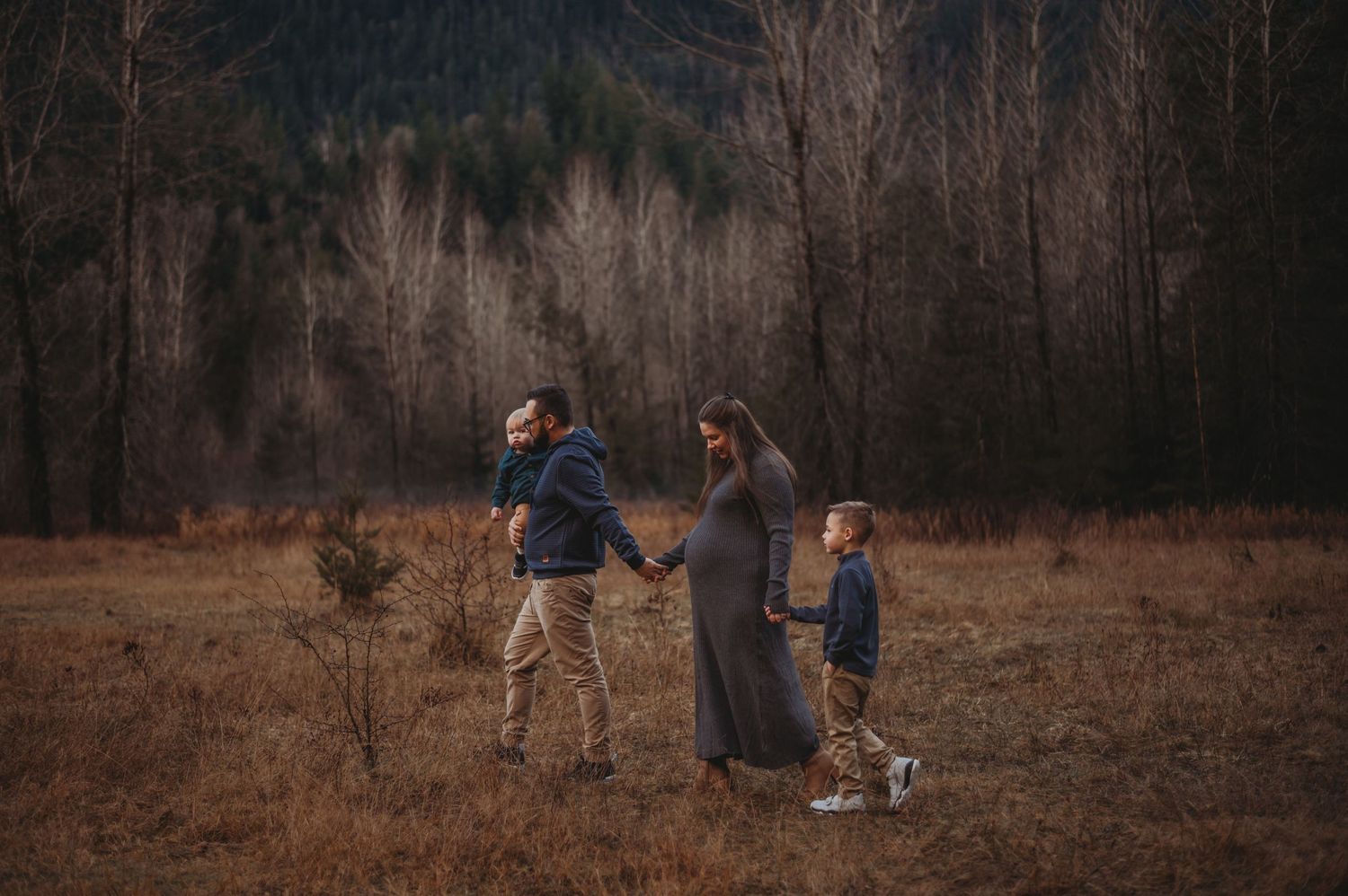 Pregnant woman in gray dress holding hands with partner and child in autumn forest clearing.