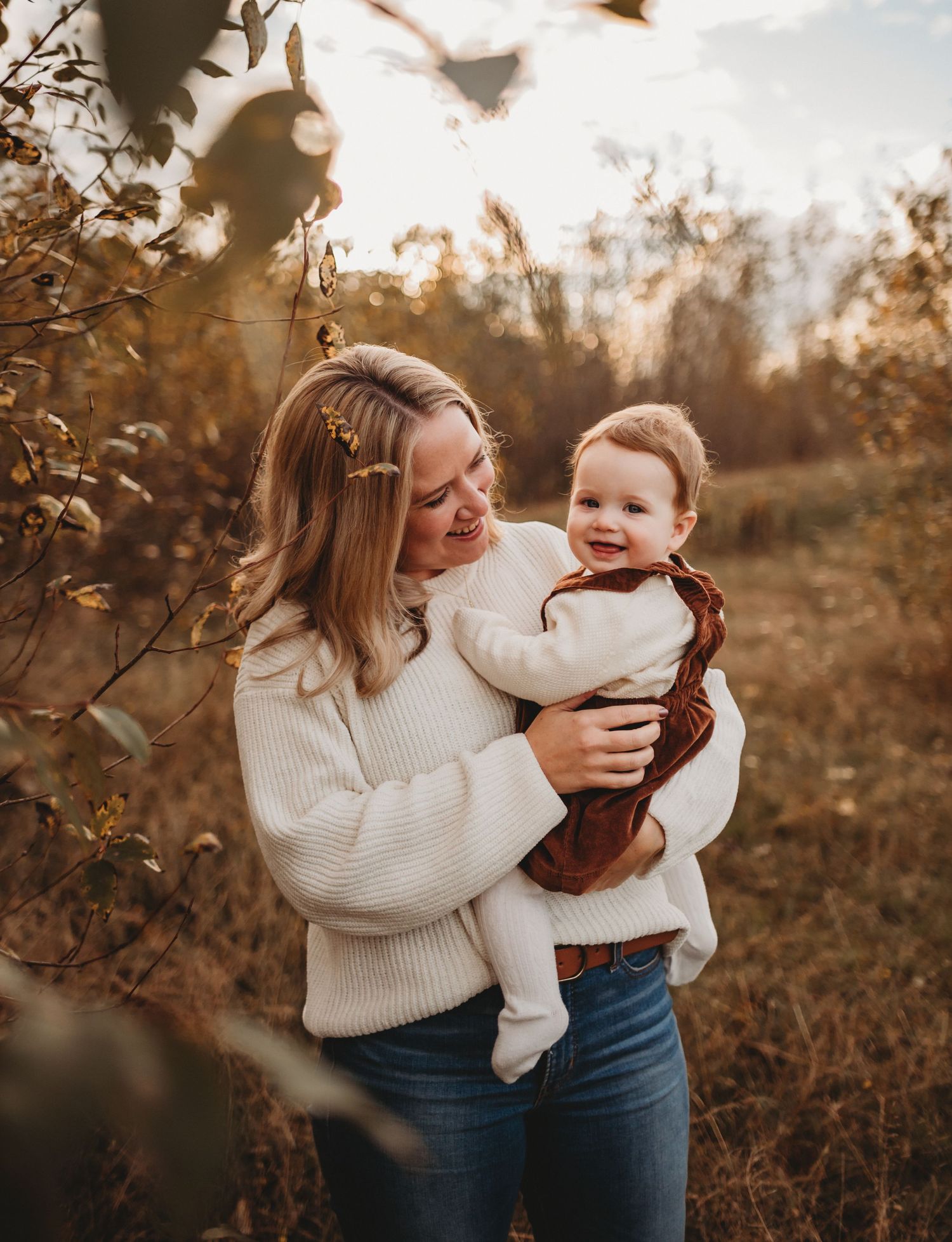 A blonde mother in a cream sweater holds her baby in a sunlit autumn field.