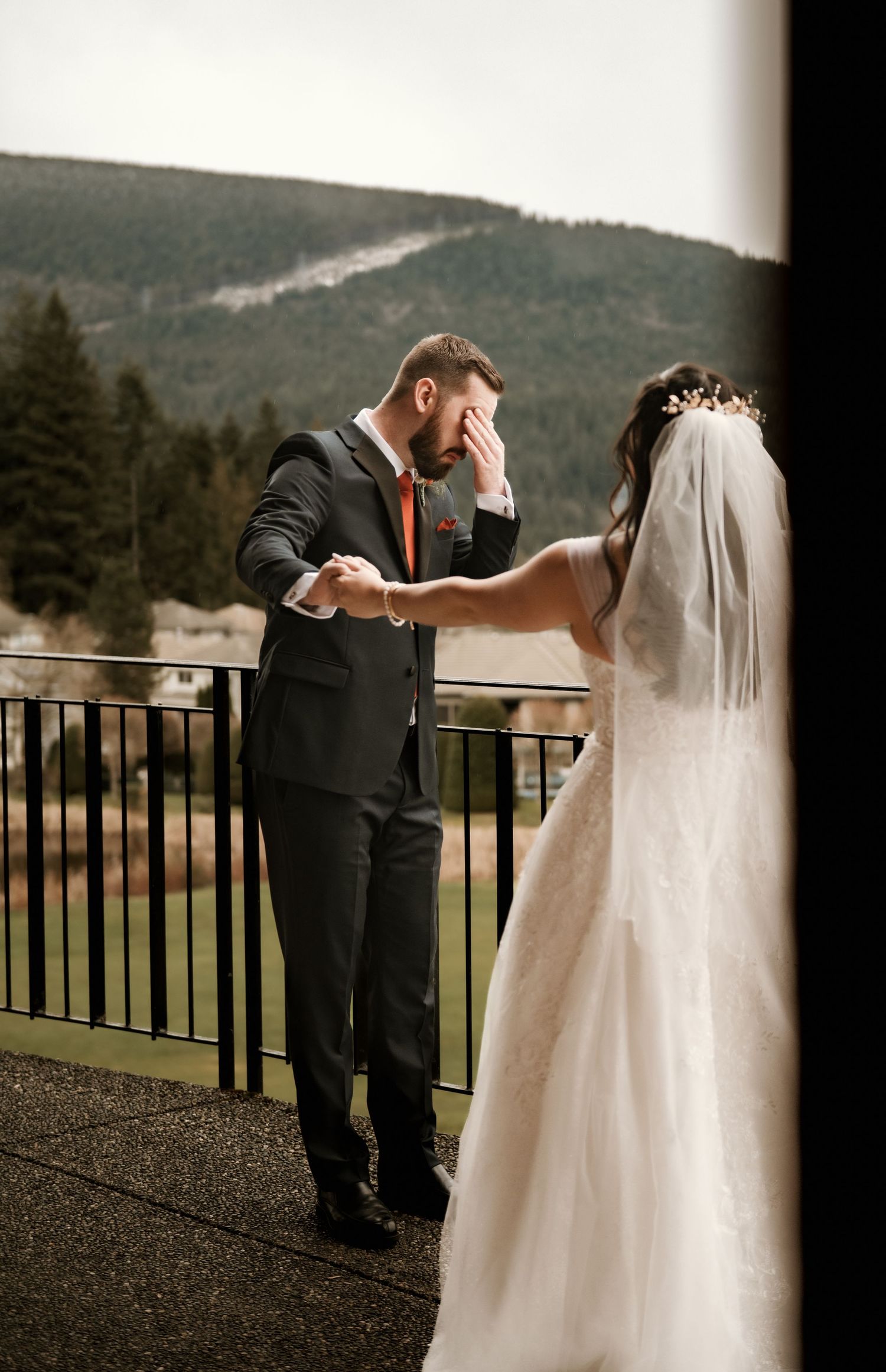 Emotional groom covering face while bride touches his hand on balcony with mountain backdrop.