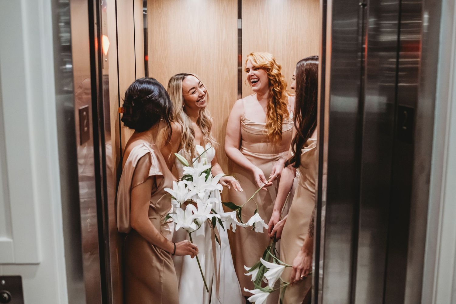 Bridesmaids in champagne gowns laugh together holding white lilies inside an elevator.