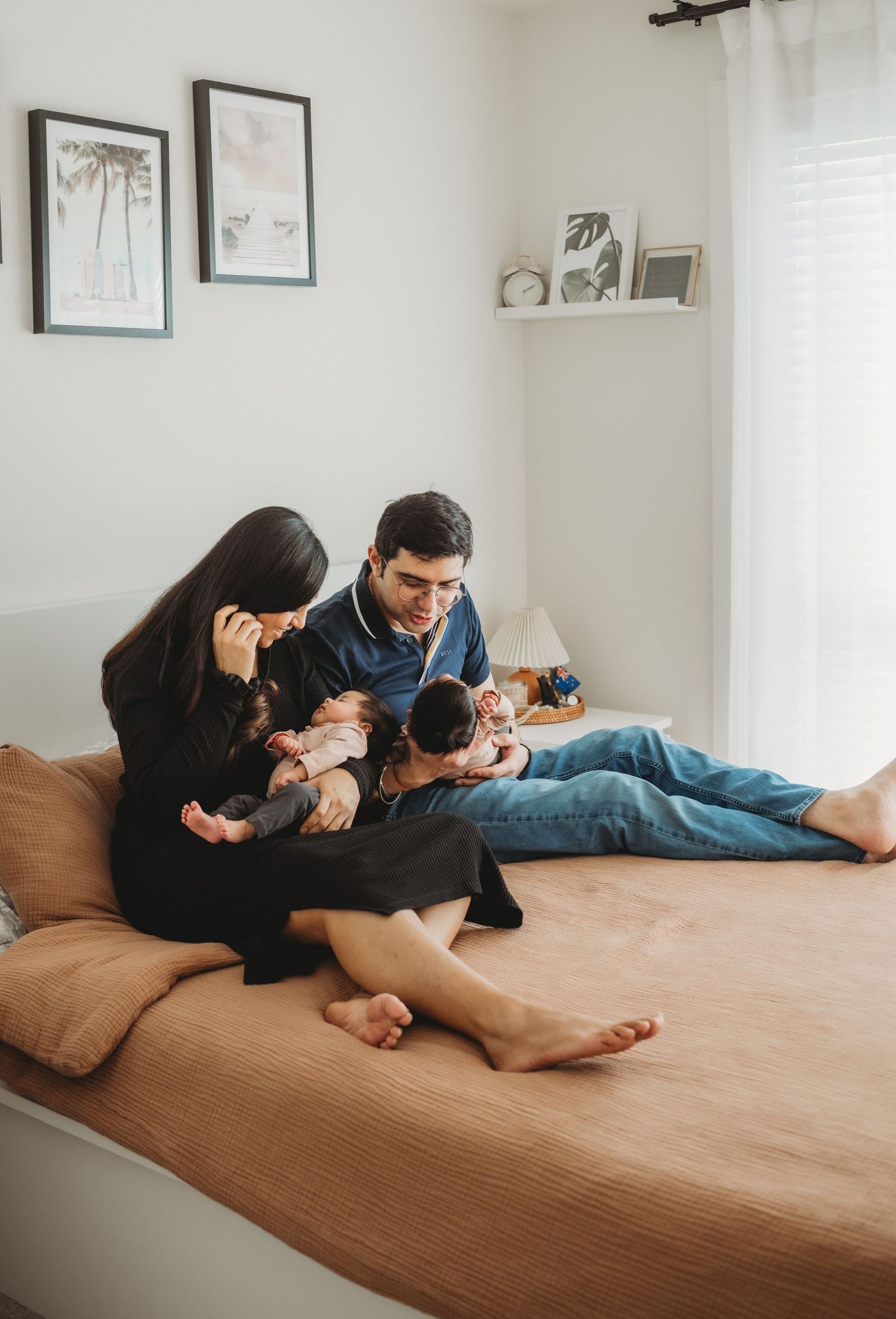 Parents relaxing on a bed holding their newborn twins in a bright cozy bedroom setting.