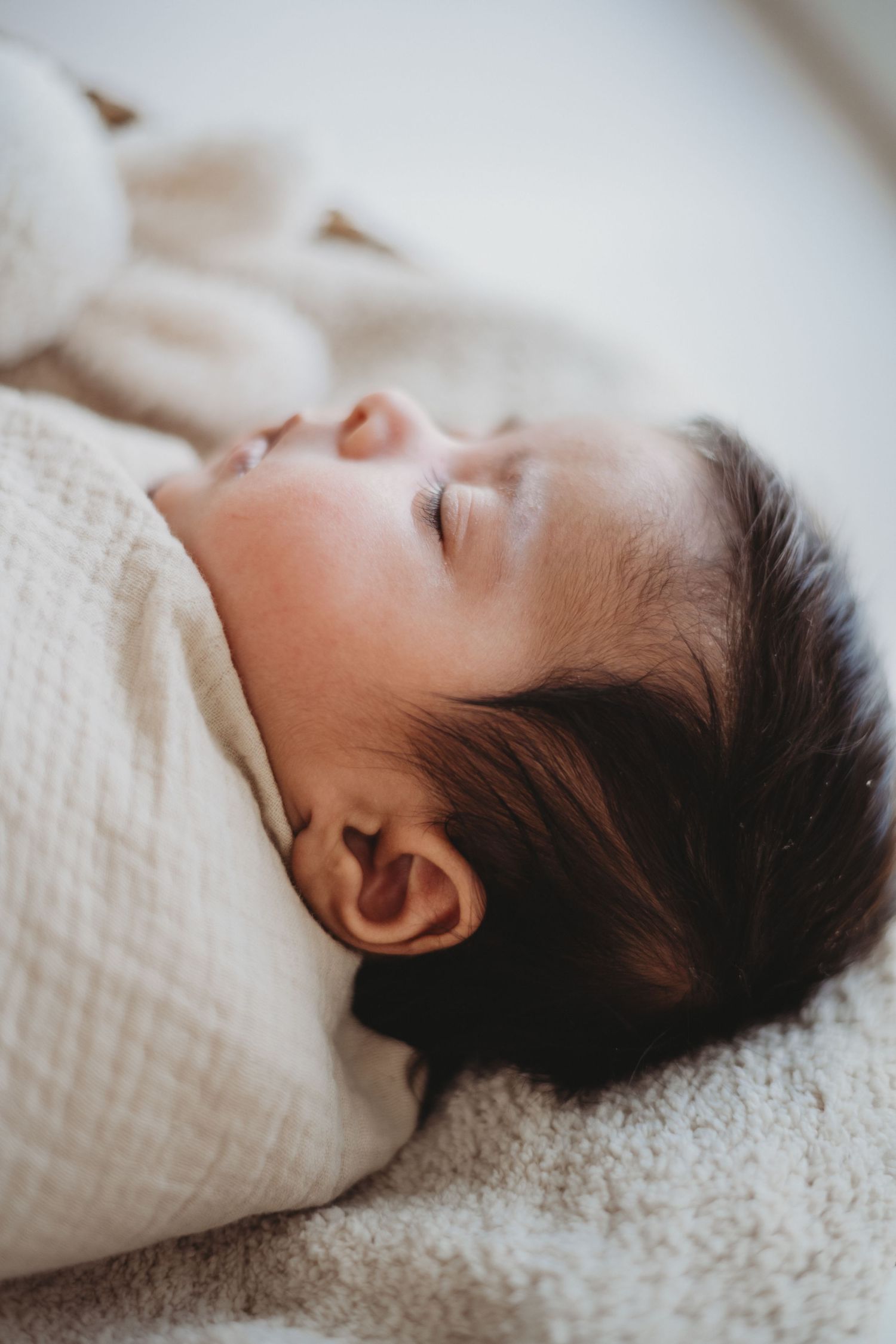 Close-up side profile of sleeping newborn baby swaddled in cream muslin blanket.