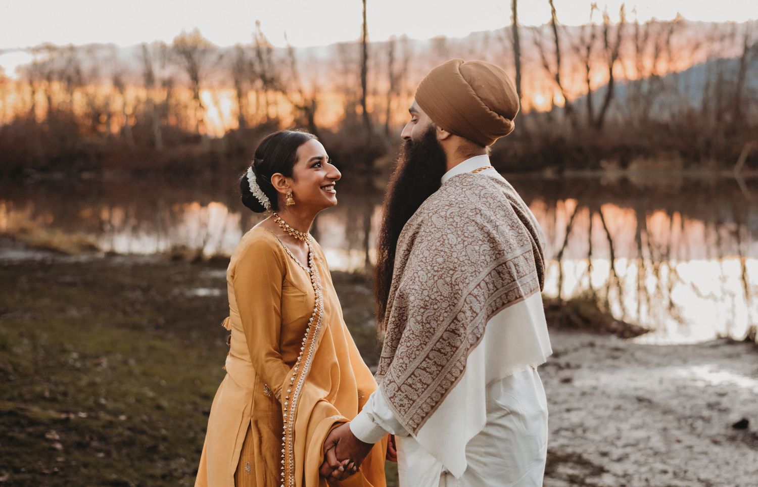 A Sikh couple in traditional attire holds hands smiling at sunset by a tranquil riverside.