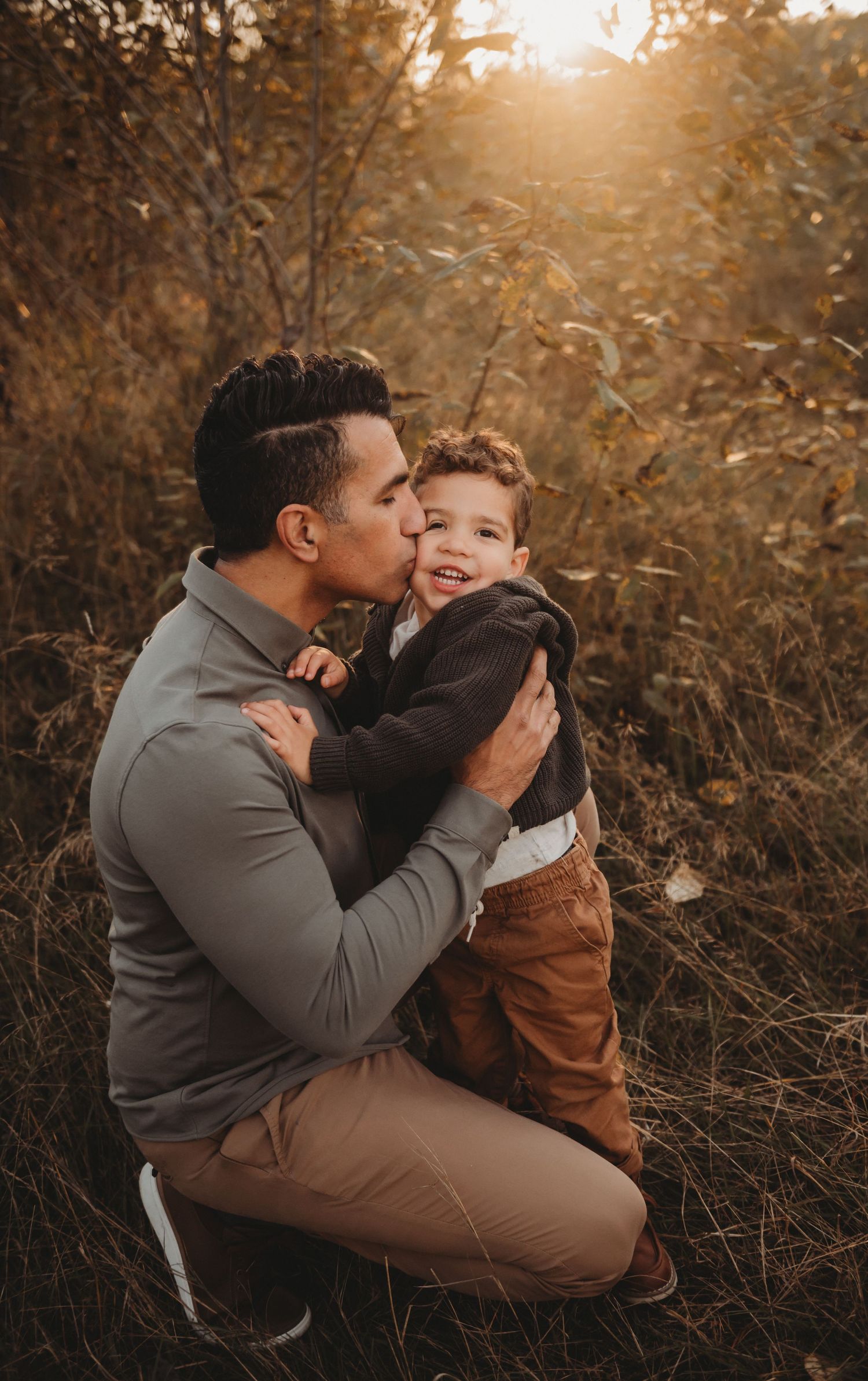 Father kissing his smiling young son on the cheek while kneeling in a golden autumn field.