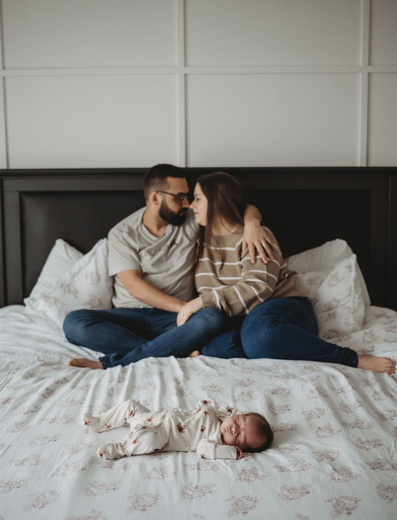 A couple sits on a bed sharing a tender moment while their newborn baby lies before them.