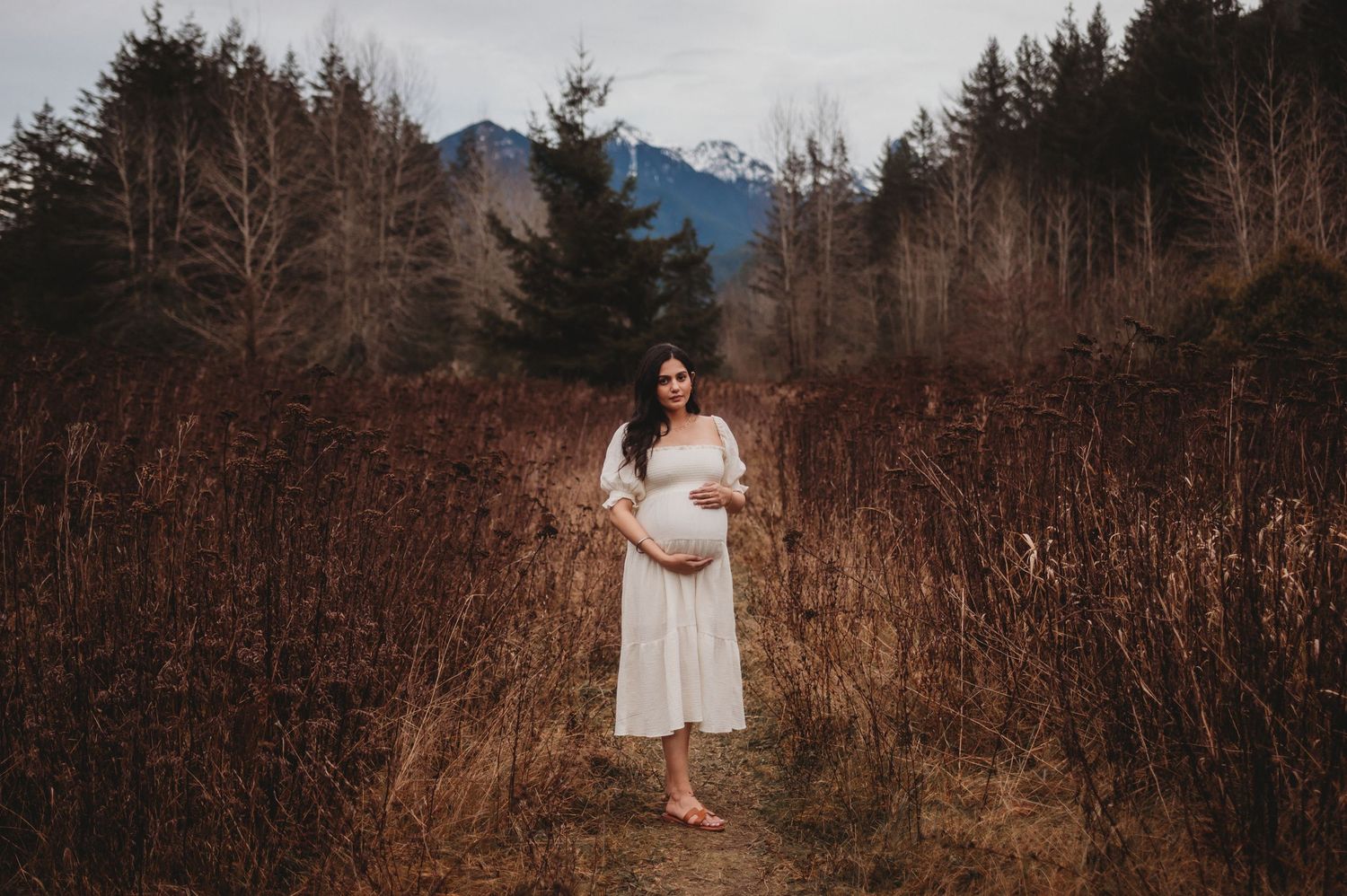Expectant mother in white dress poses among tall dried grasses with mountain scenery behind her.