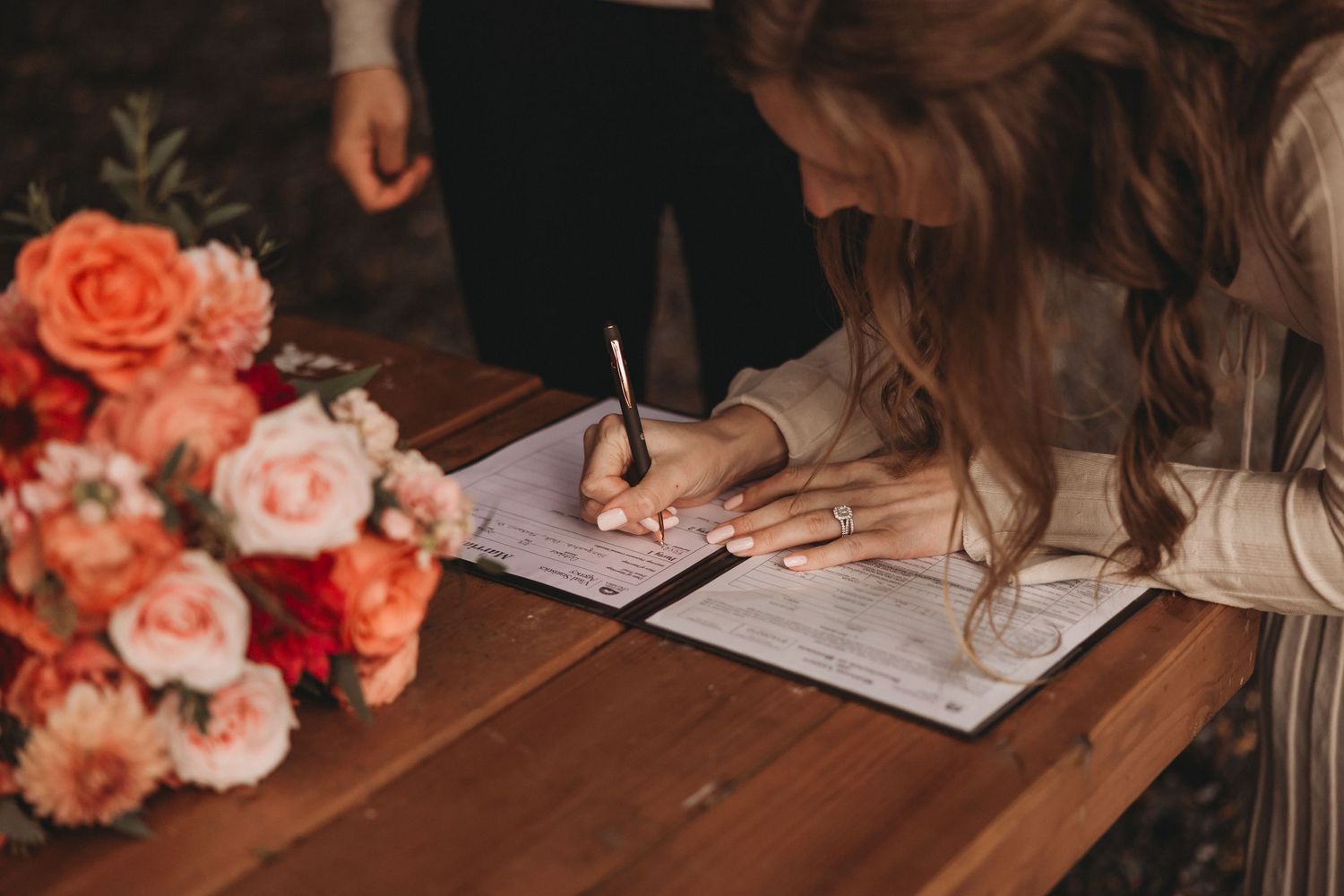 Bride signing wedding certificate at wooden table beside vibrant orange and pink floral arrangement.