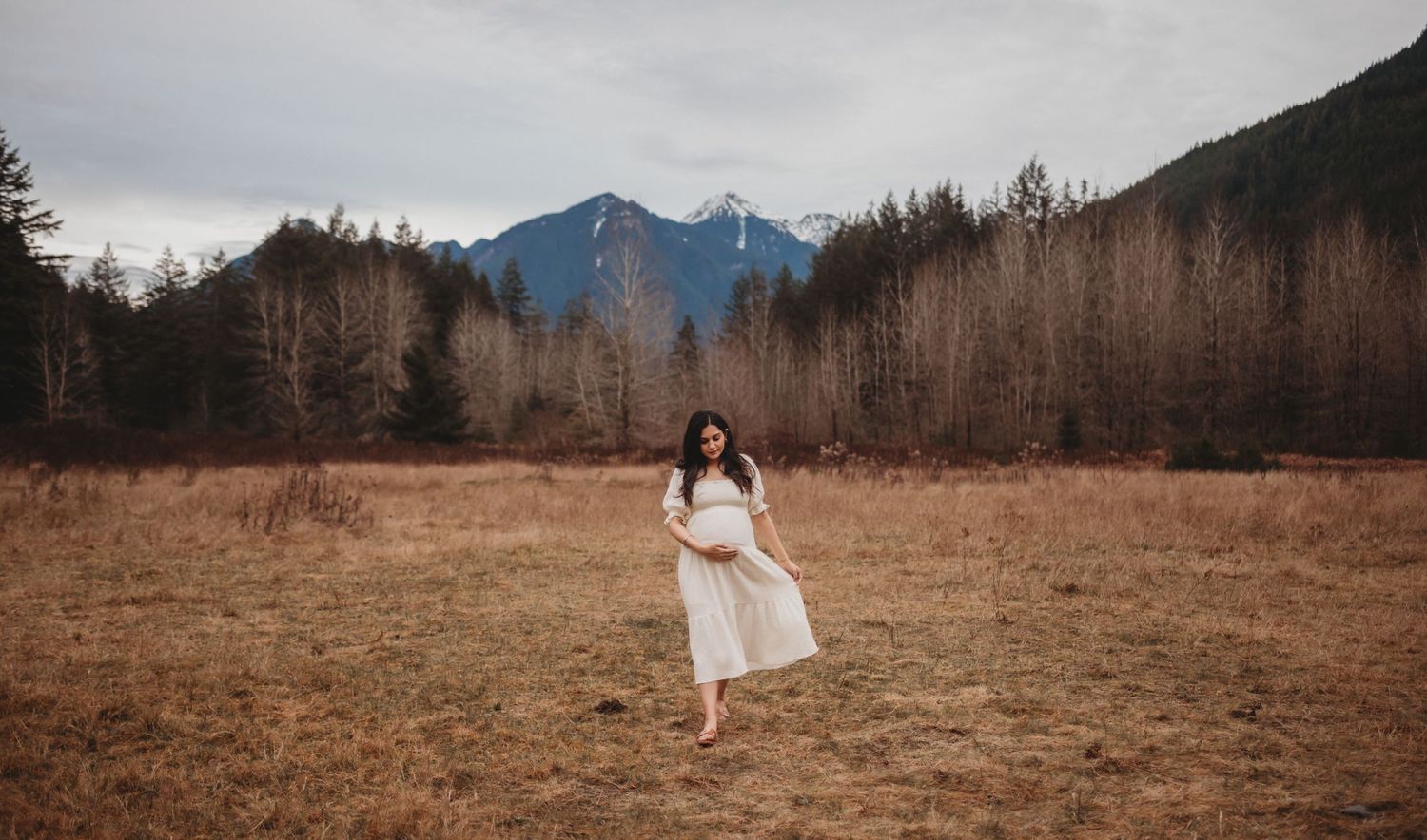 Pregnant woman in white dress walks through golden meadow with snow-capped mountains in background.
