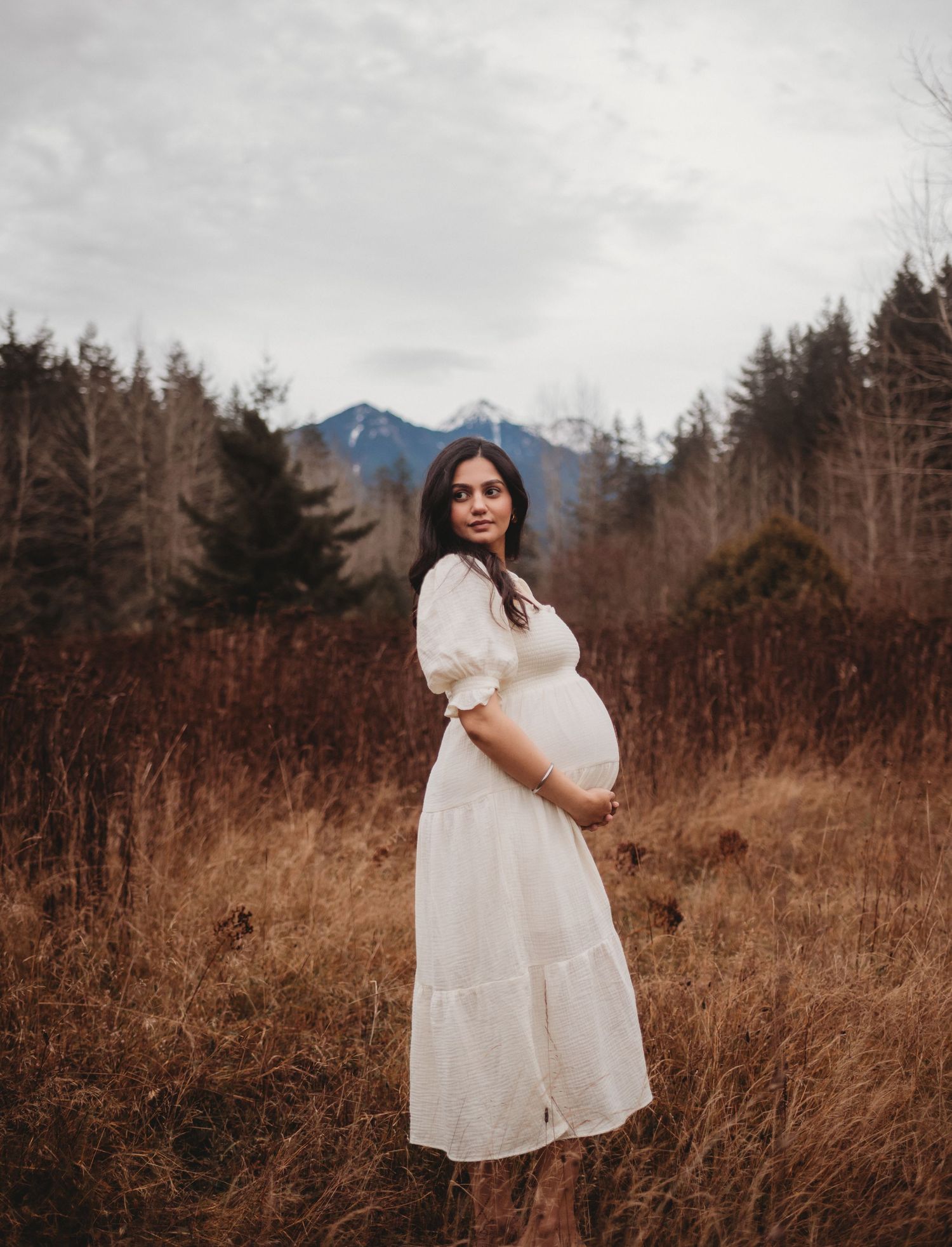 Smiling pregnant woman in white dress looking over shoulder in rustic field with snow-capped mountains.