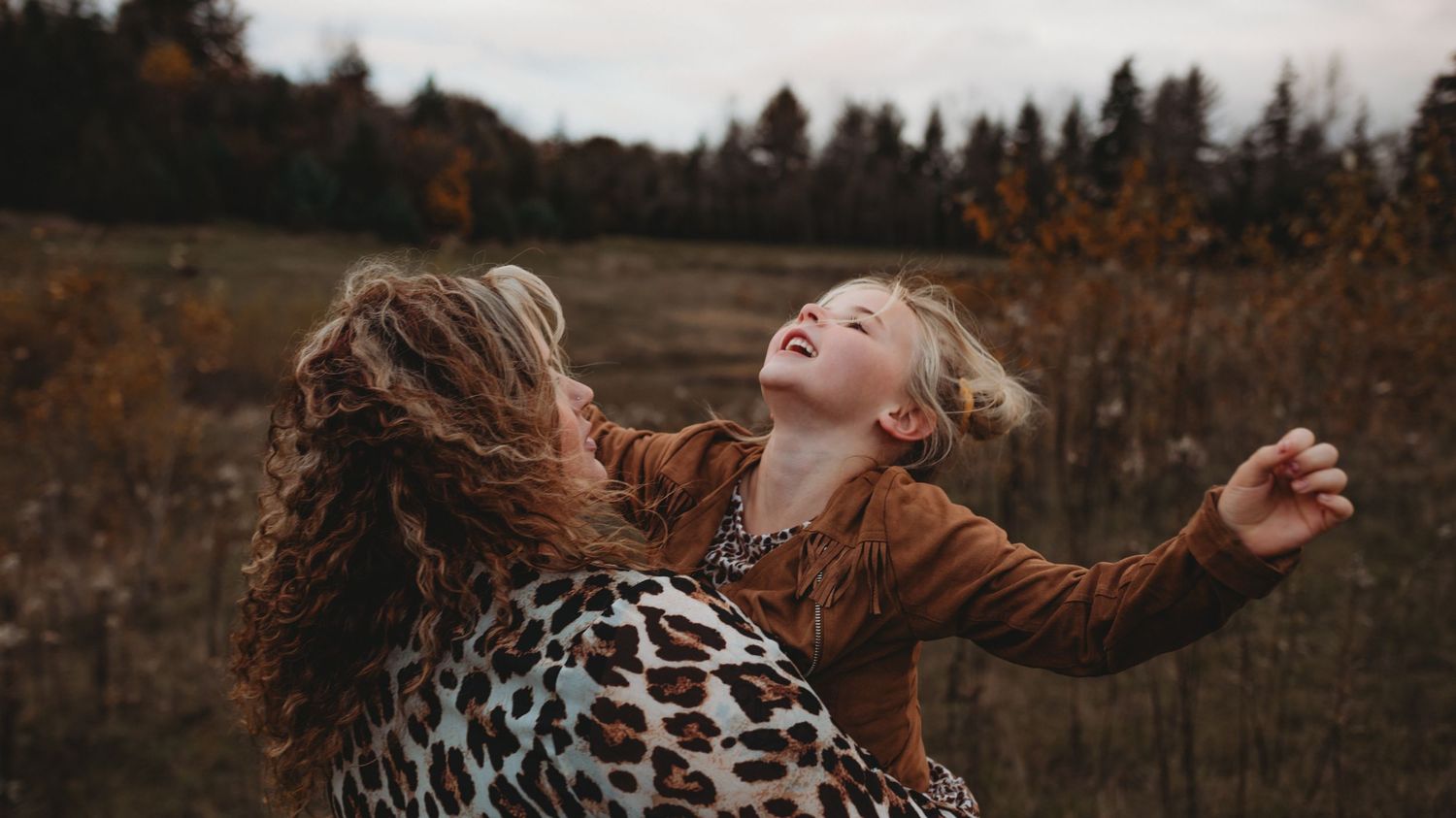 Woman in leopard print top lifting laughing young girl in brown jacket in autumn field.