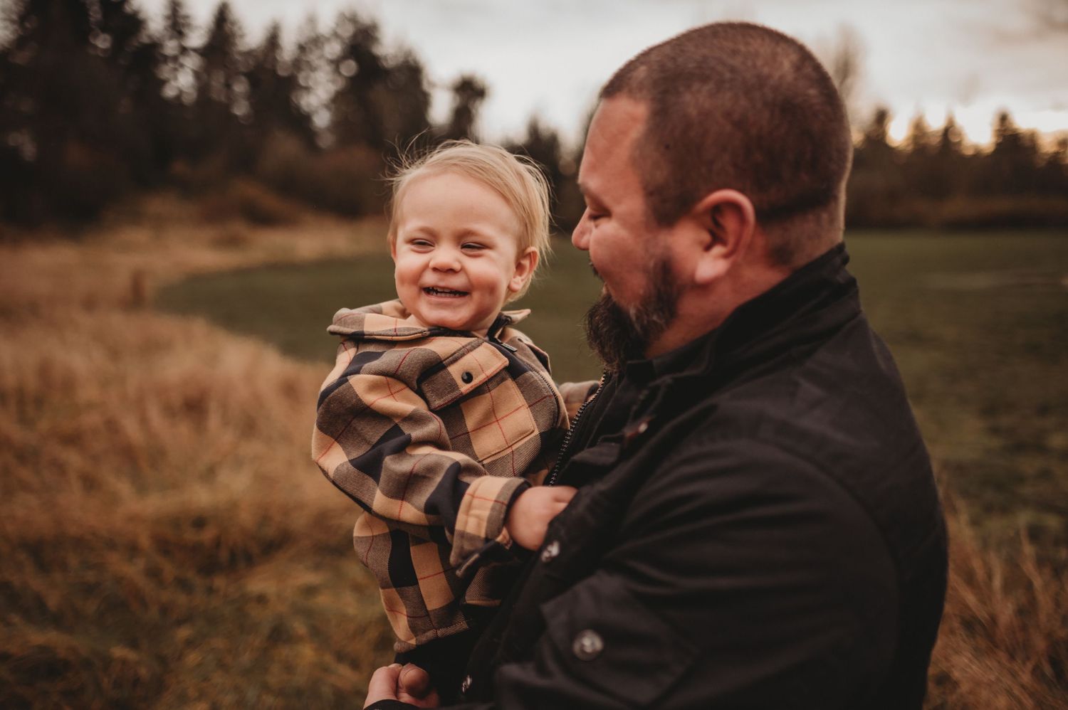 Father laughing with smiling toddler son in plaid shirt outdoors.
