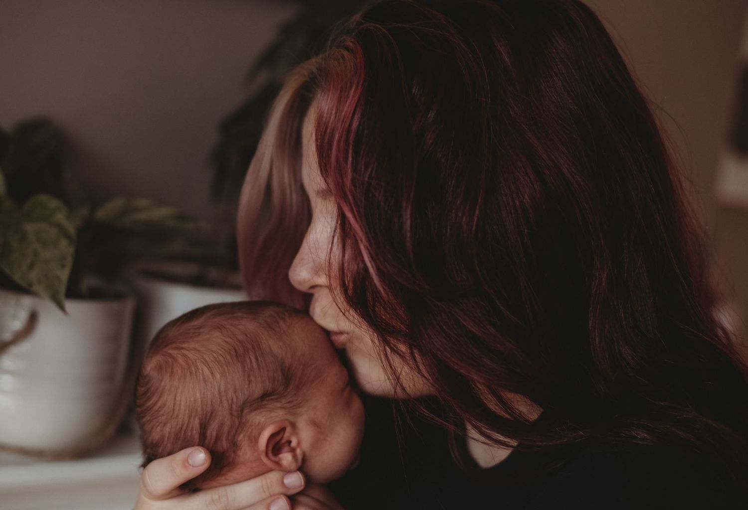 Red-haired mother gently kissing her newborn baby in a softly lit room.