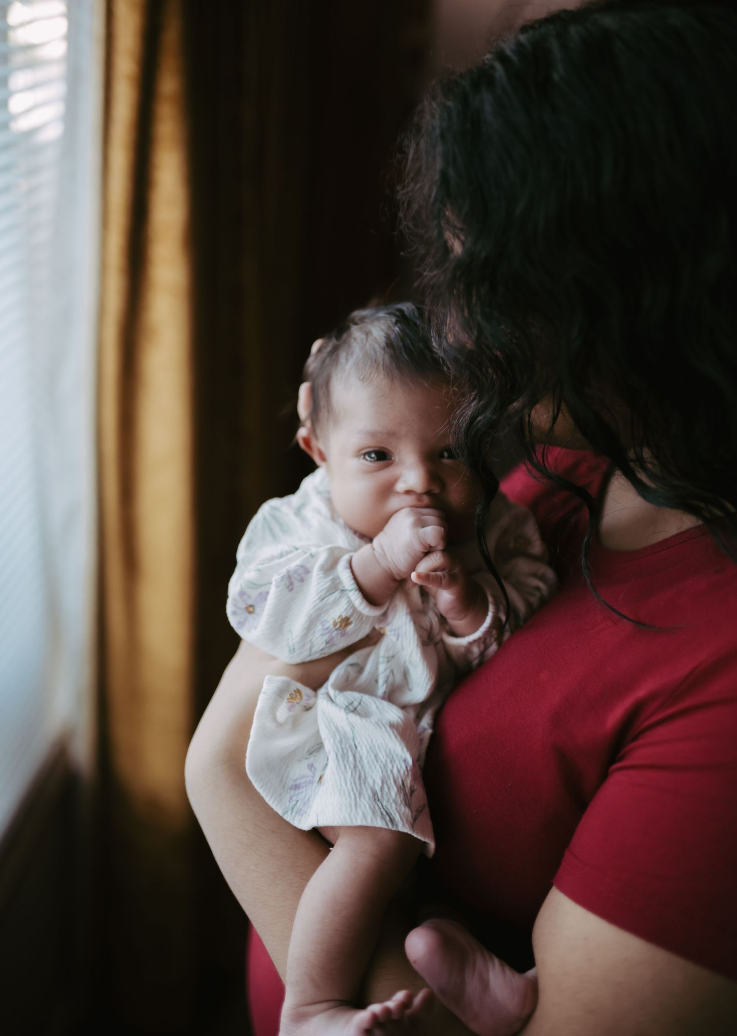 Mother in red top cradling alert newborn baby in white floral dress near window.