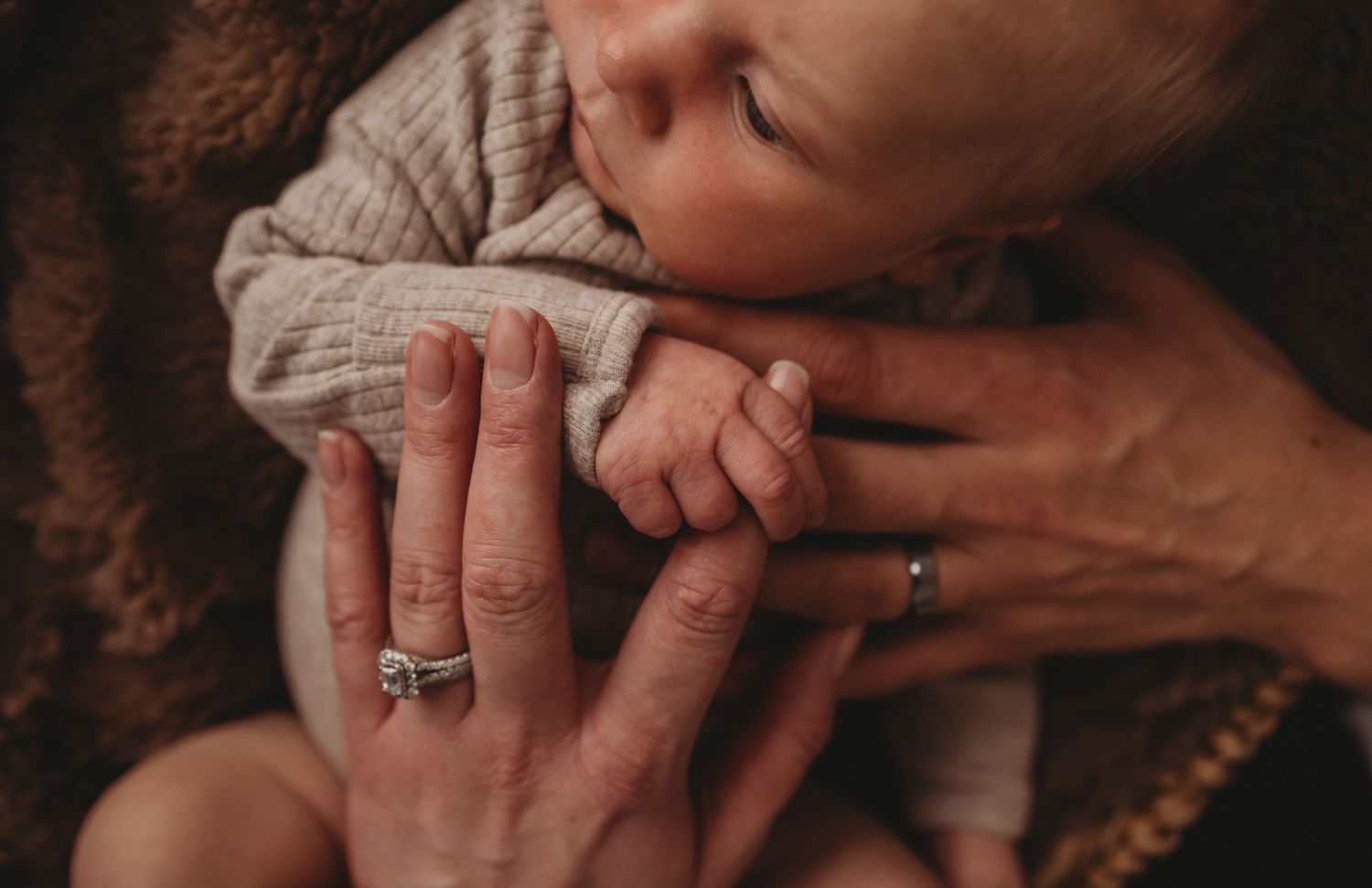 Close-up of parents hands gently holding sleeping newborns tiny fist, both wearing rings.