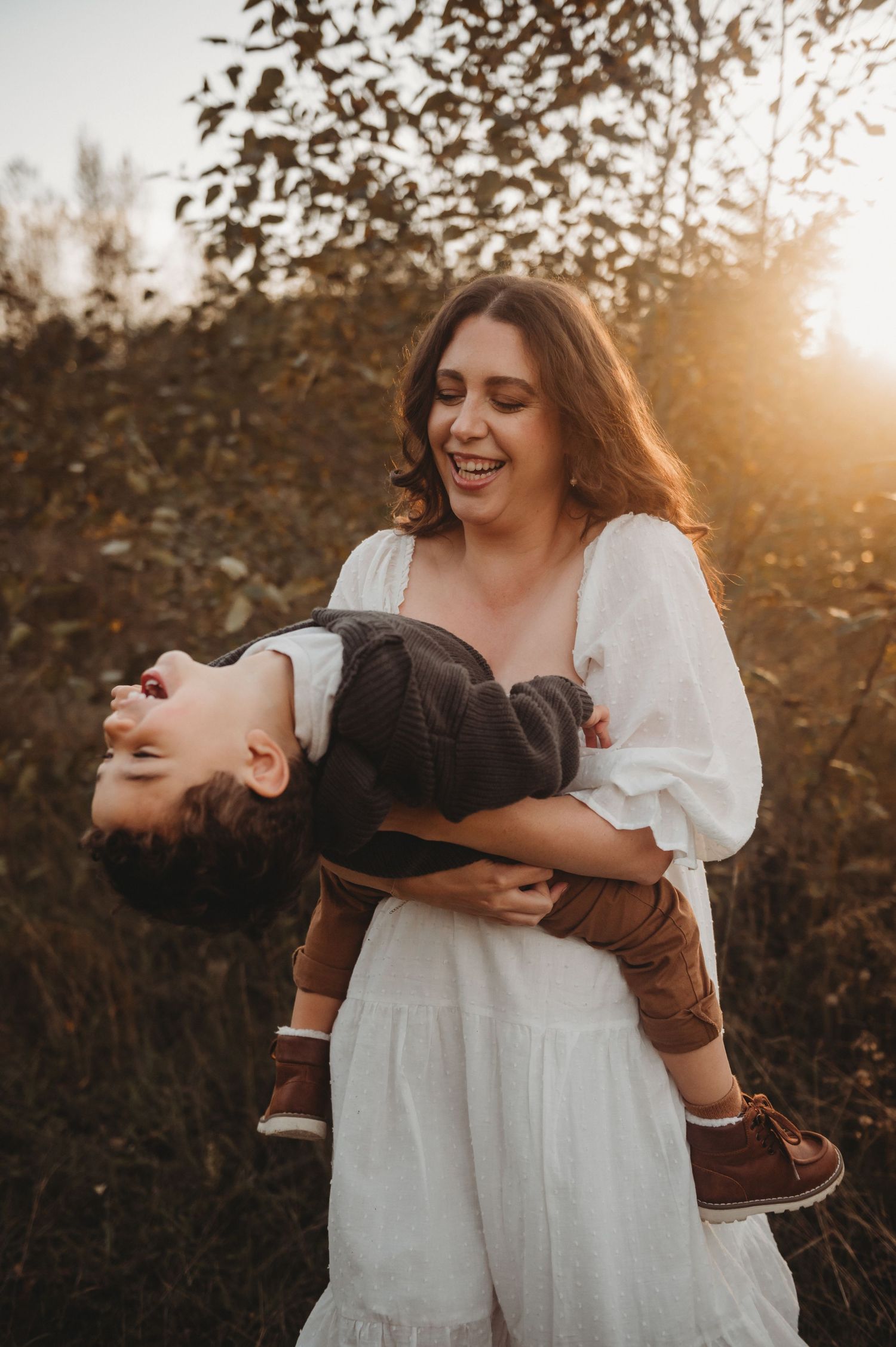 Joyful mother laughing while holding her son upside down in a glowing autumn outdoor setting.