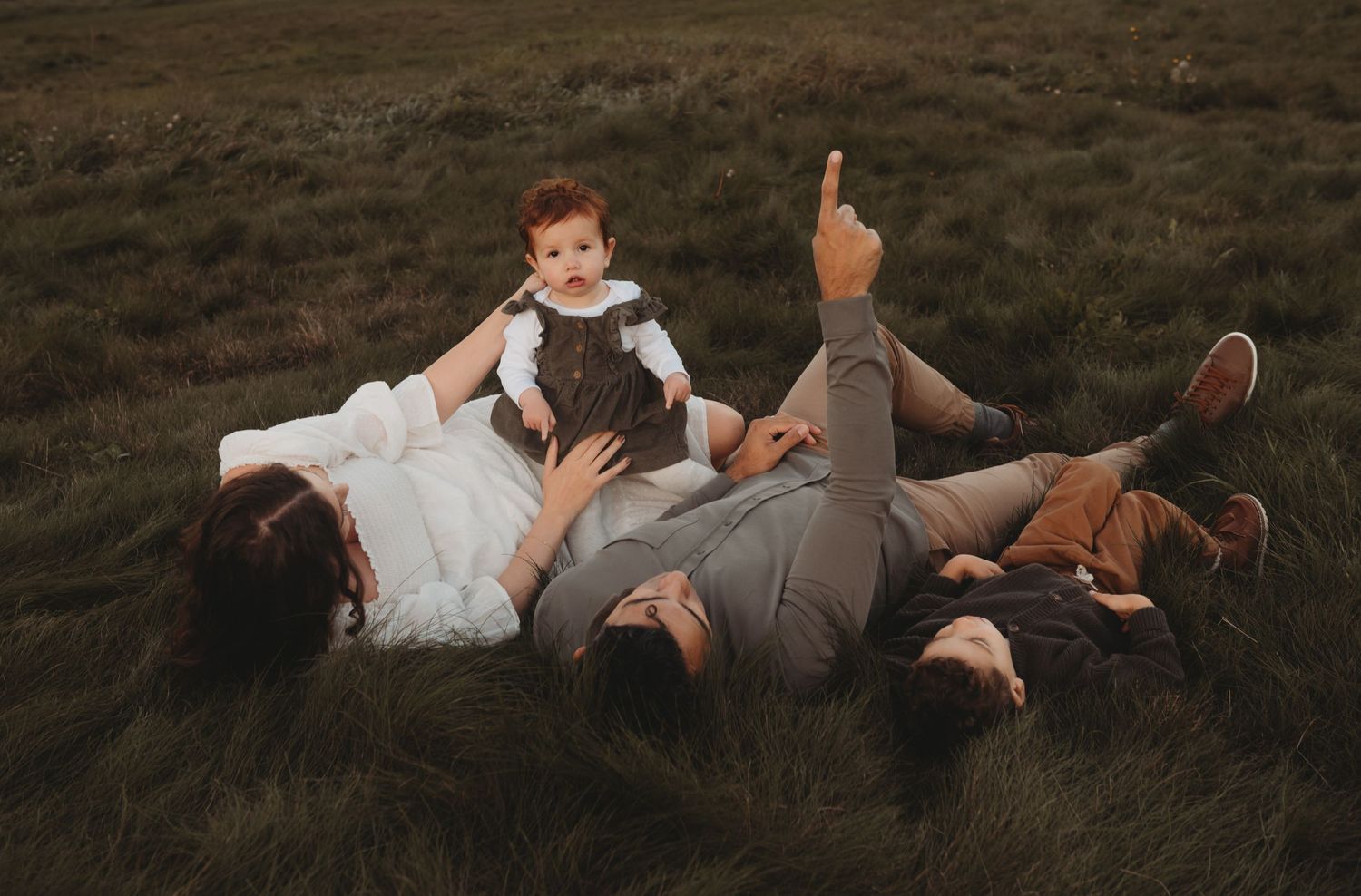 A family relaxes on lush grass, with a baby sitting up while parents and older child lay down together.