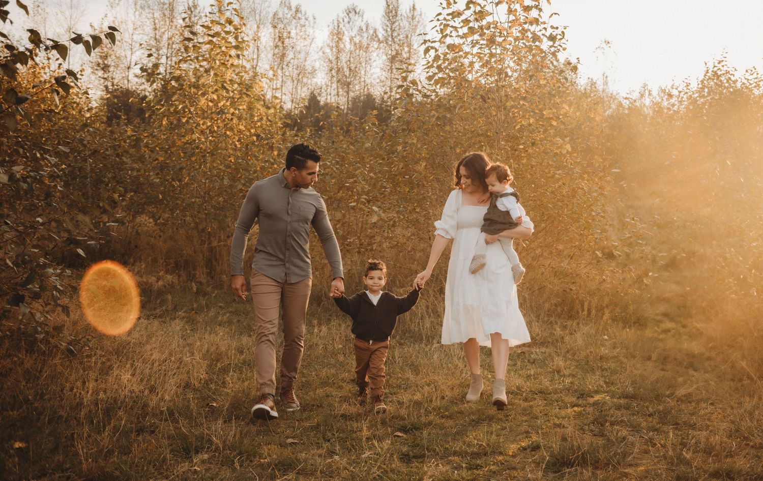 A couple walks with two young children through a glowing autumn field at golden hour.