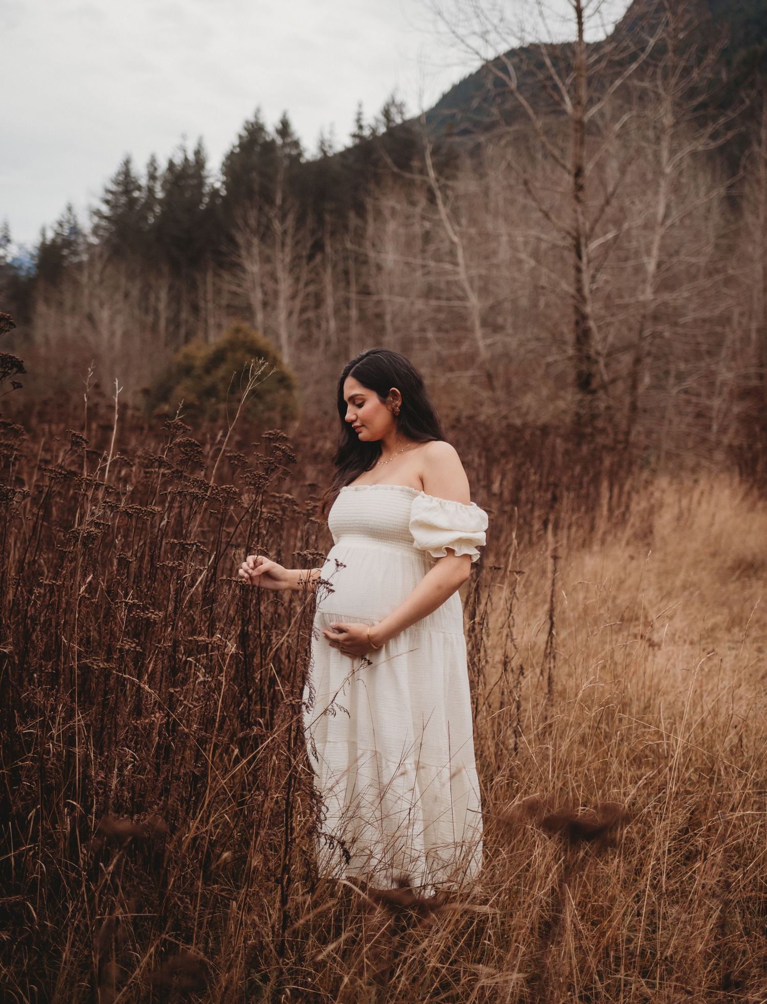 Pregnant woman in white off-shoulder dress standing in golden field with mountains and bare trees.