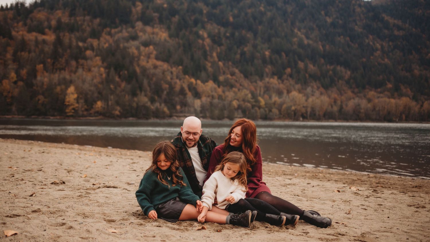 A family of four sits together on a lakeside beach surrounded by fall mountain scenery.