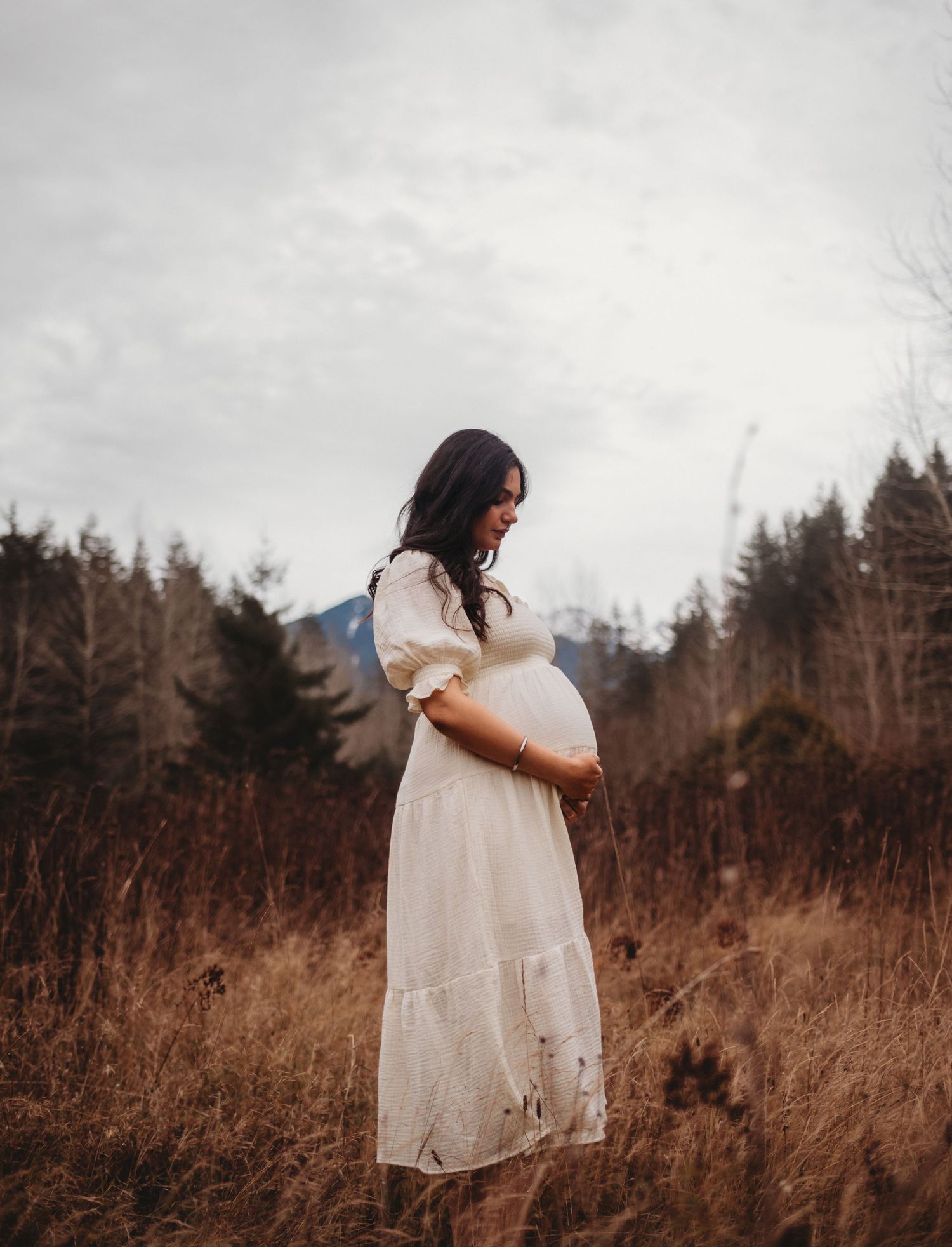 Side profile of pregnant woman in white dress gently holding baby bump in golden field near forest.