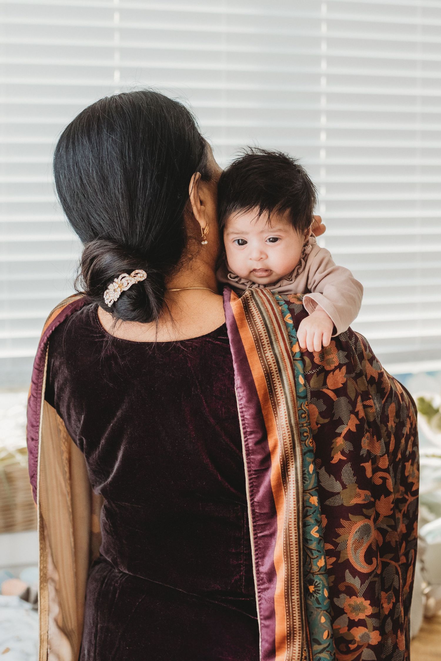 Woman in purple velvet saree holding a curious baby on her shoulder near window blinds.