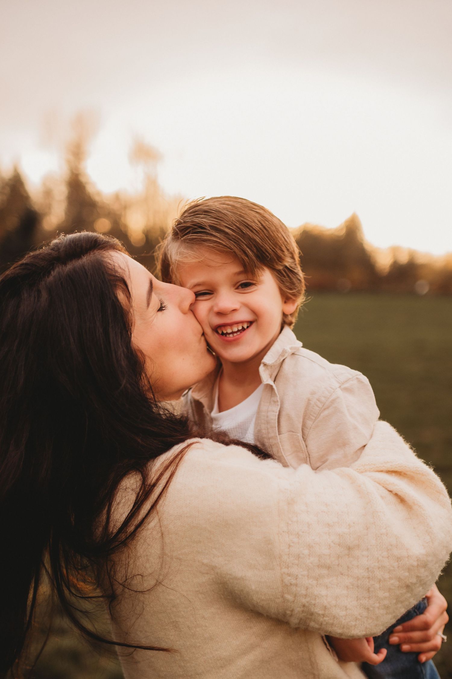 A mother kisses her laughing young boy on the cheek during a golden hour outdoor photo session.