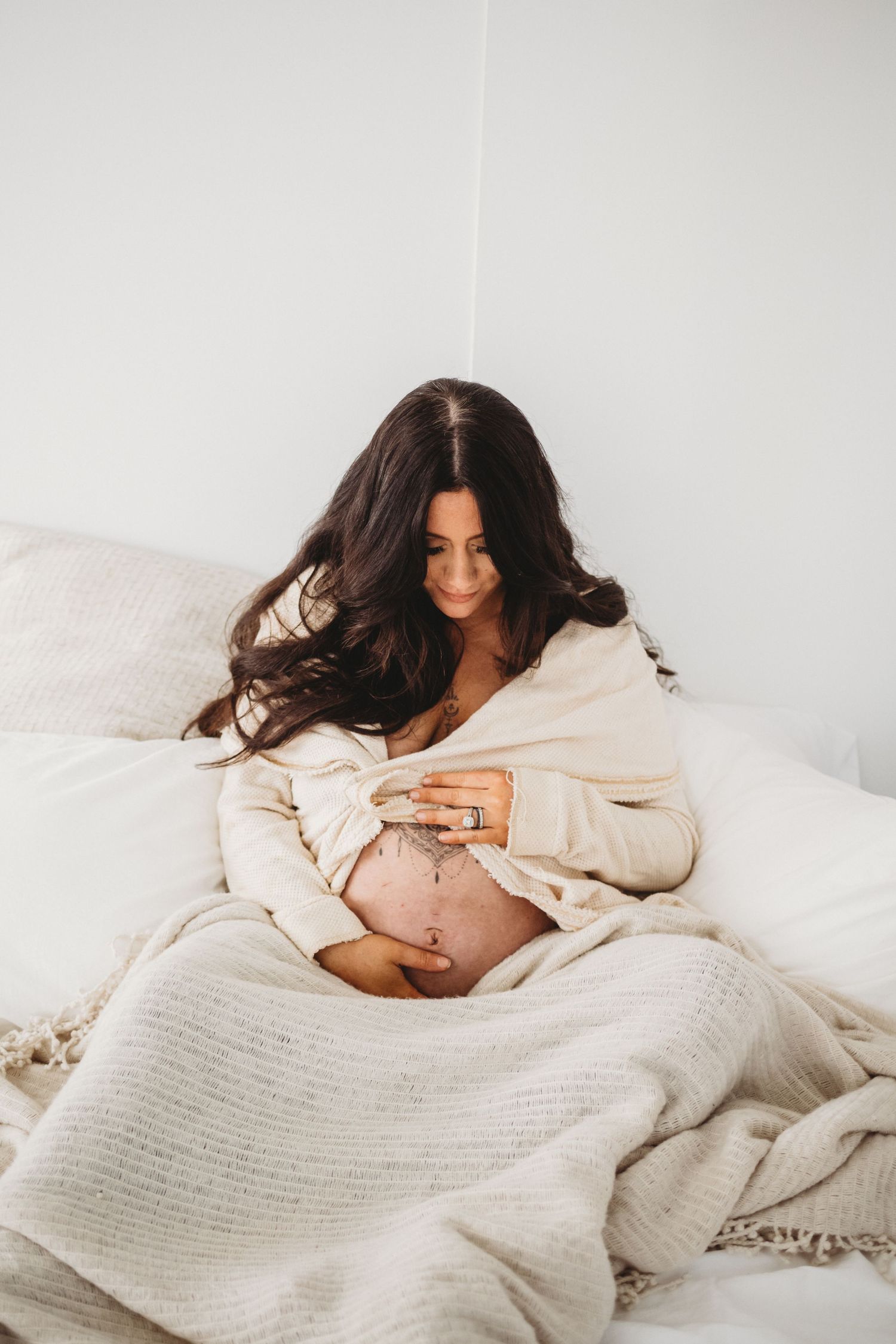 Pregnant woman in cream wrap gazing down at tattooed baby bump on bed.