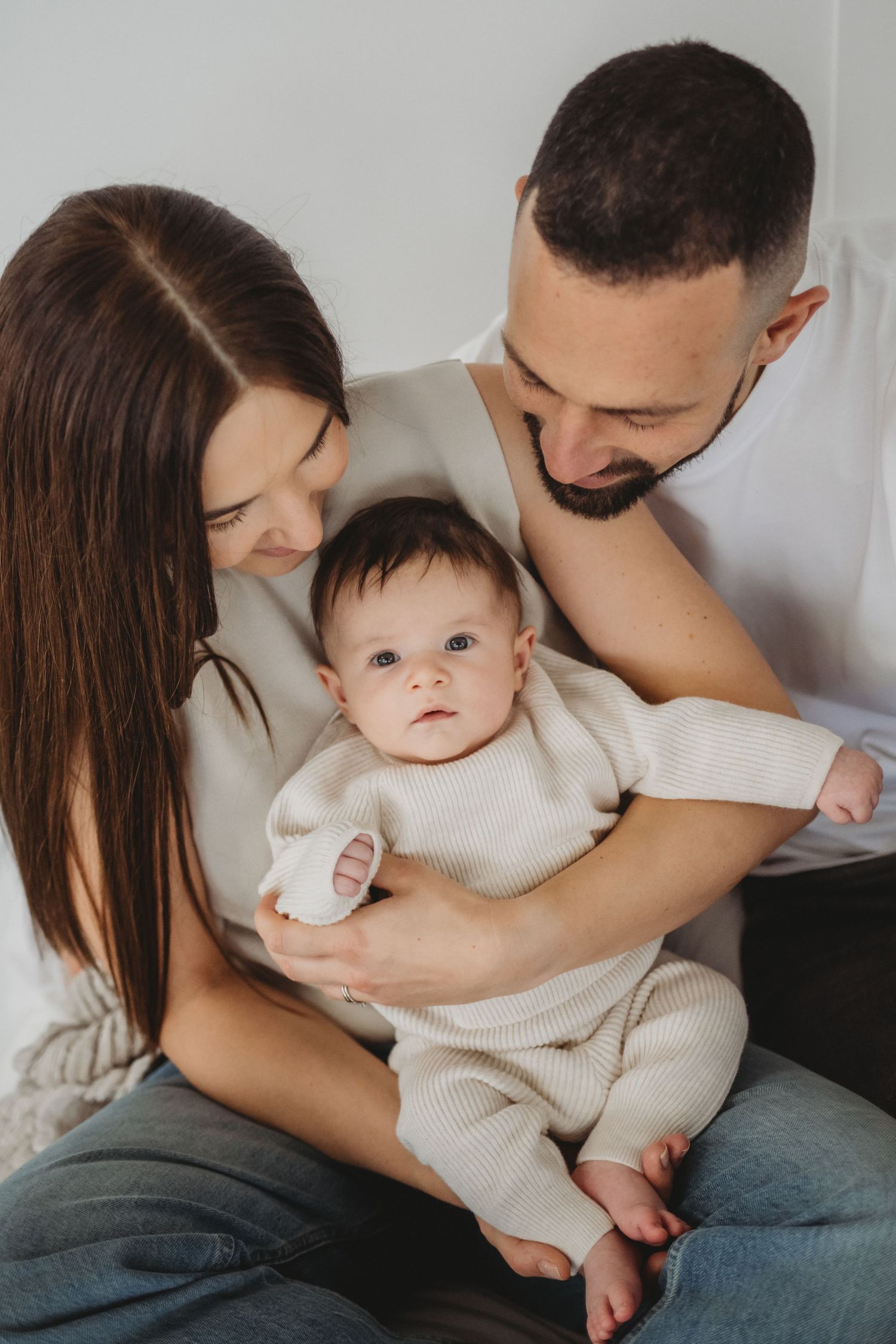Young couple in white outfits cradling their newborn baby in cream knit romper.