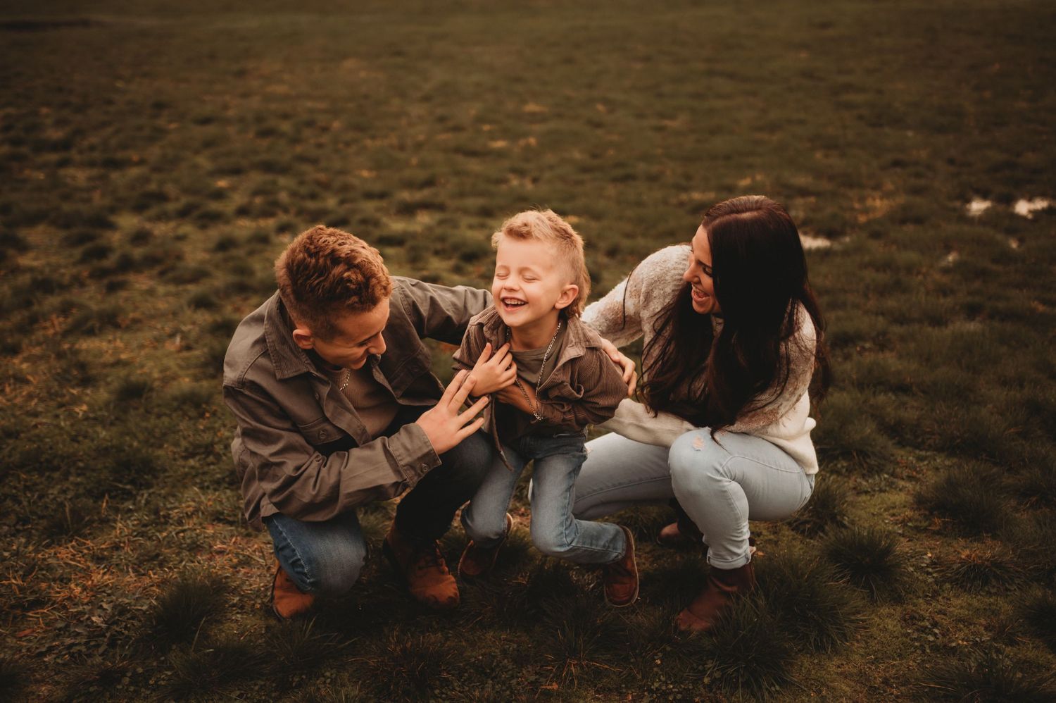 Two parents crouching and tickling their laughing toddler on a grassy field.