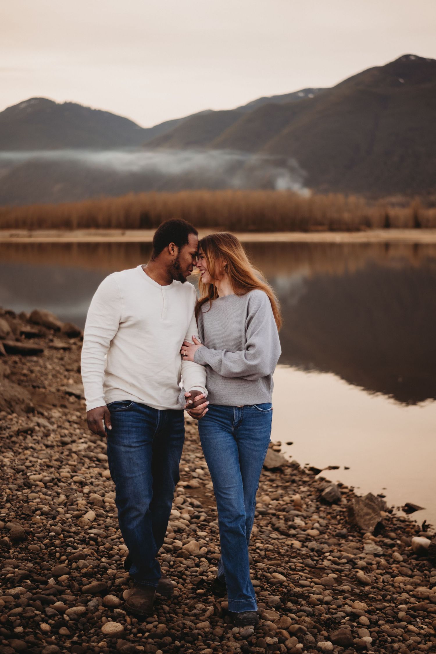 Interracial couple walking hand in hand along a rocky lakeside with misty mountains at dusk.
