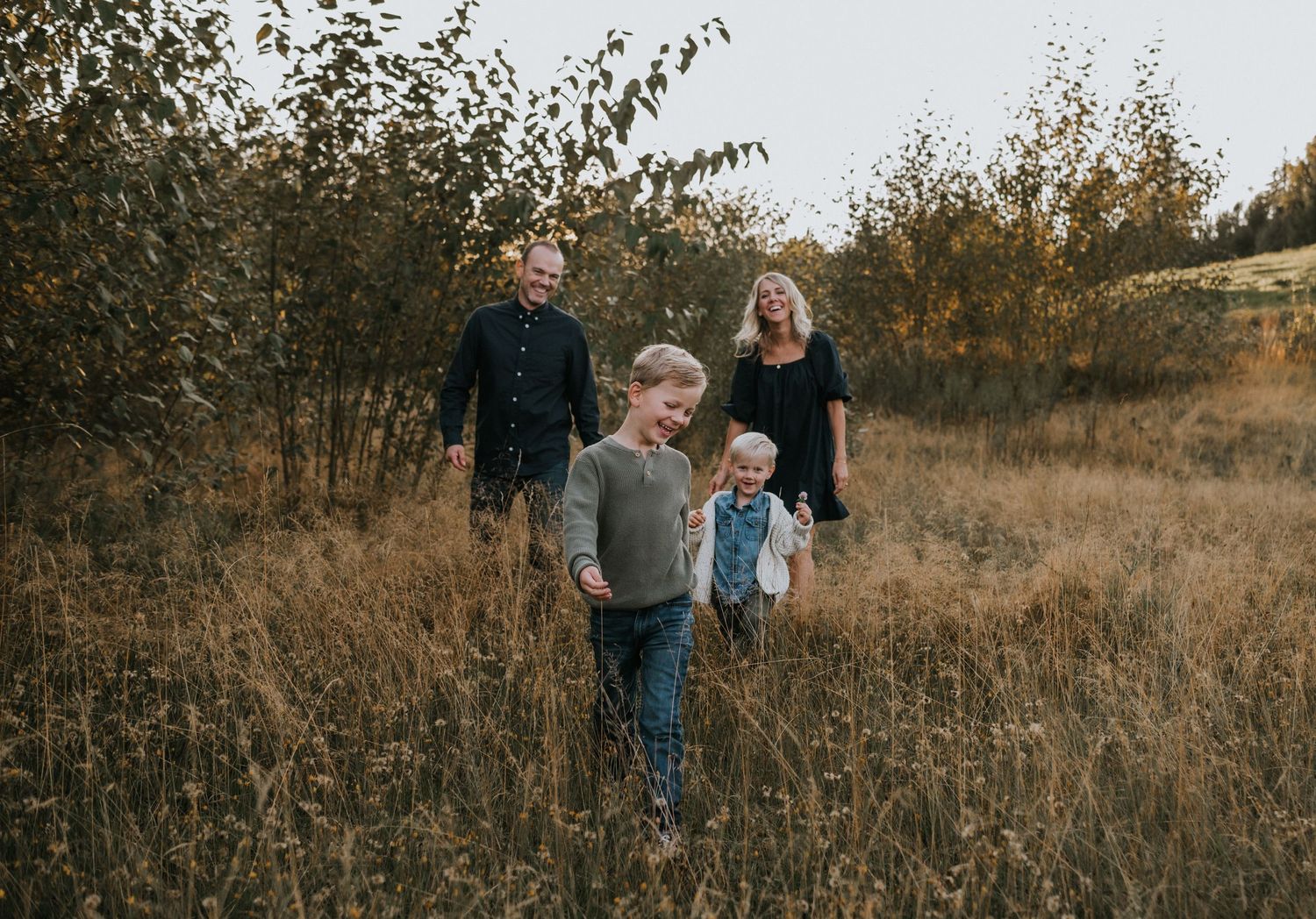 Family of four walking through golden autumn meadow with tall grasses and trees.