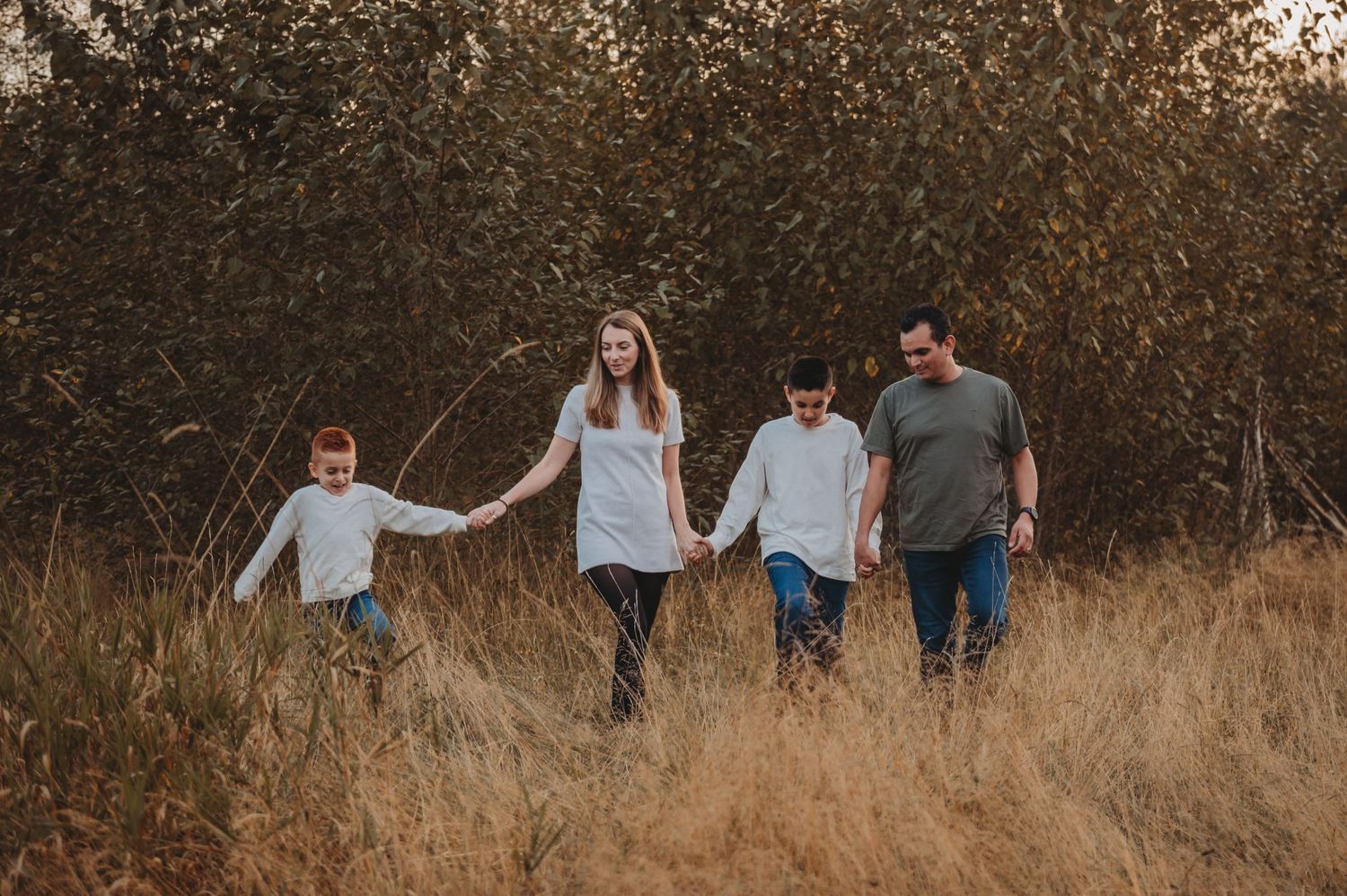Family of four walking hand-in-hand through golden autumn field with trees in background.