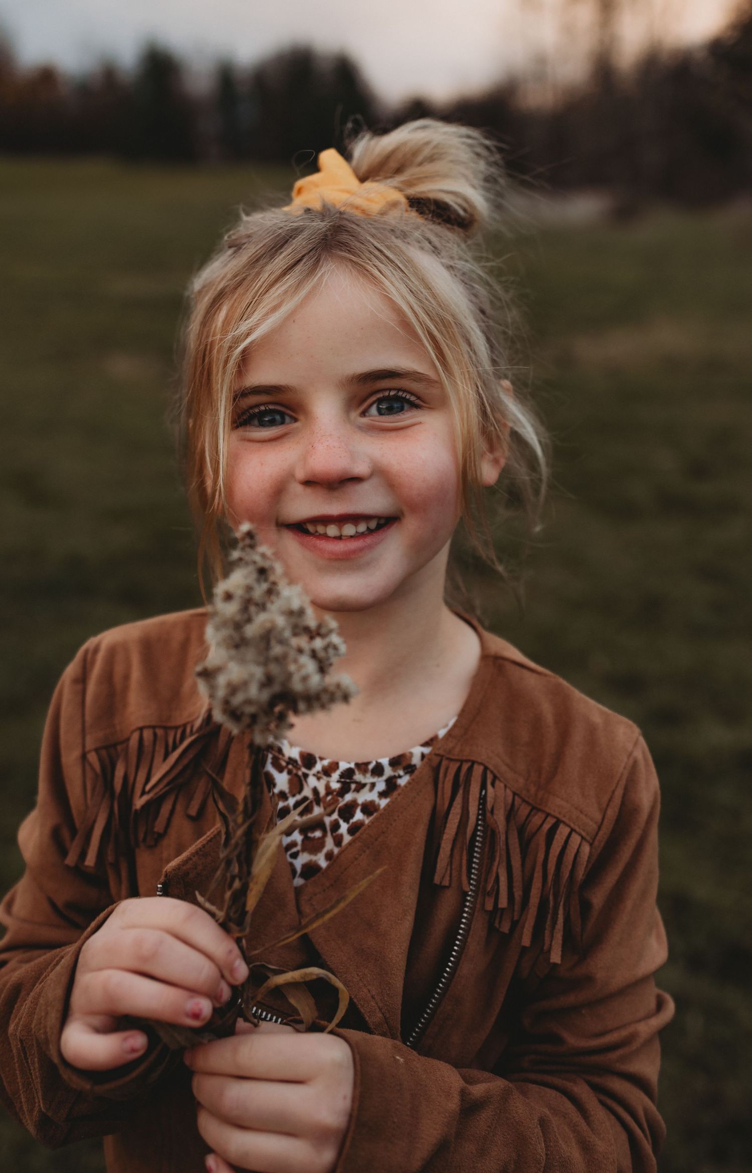 A smiling blonde girl with a yellow bow holds a dried wildflower in an outdoor autumn setting.