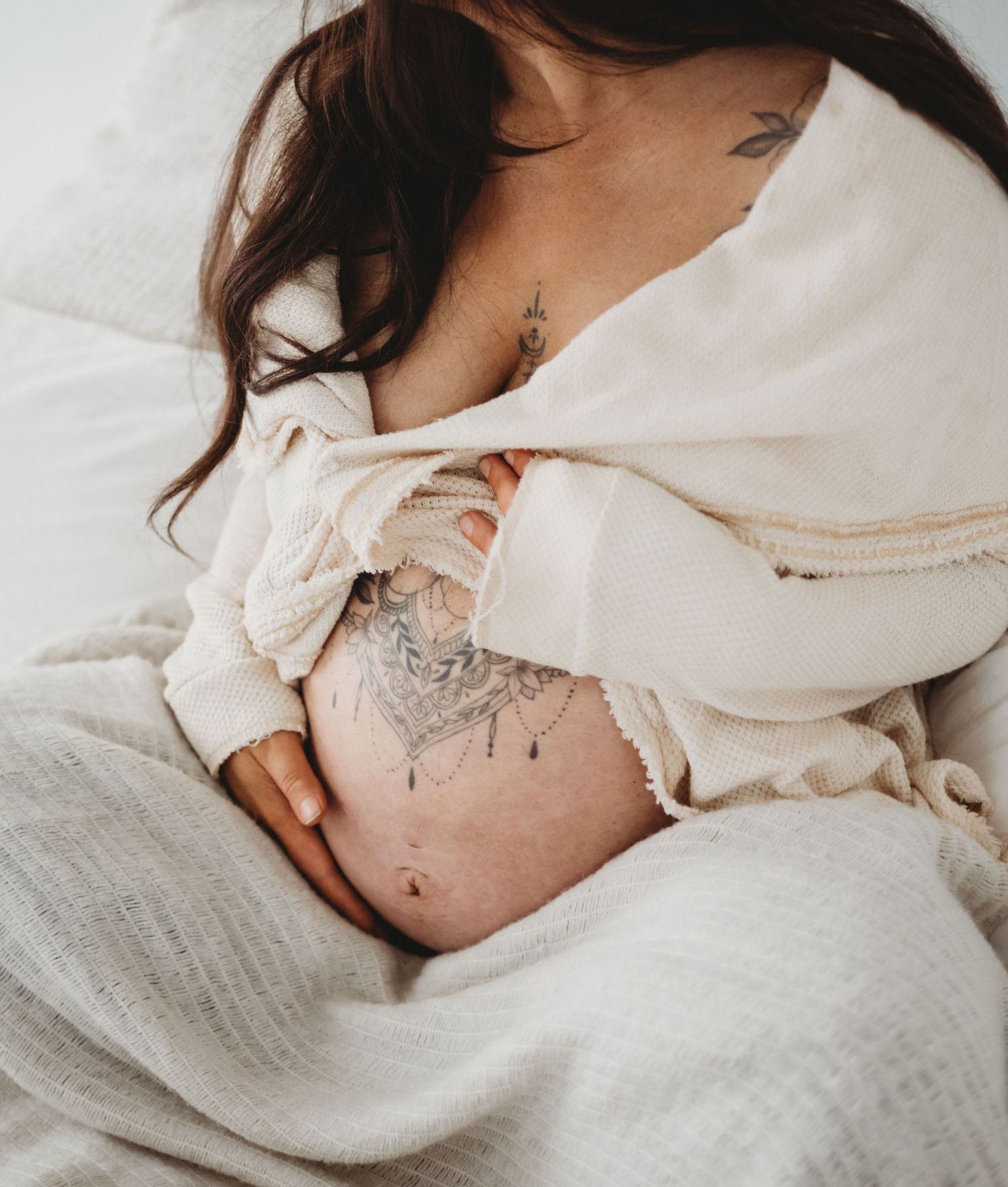 Pregnant woman with floral tattoos on belly resting on white bedding holding baby bump.