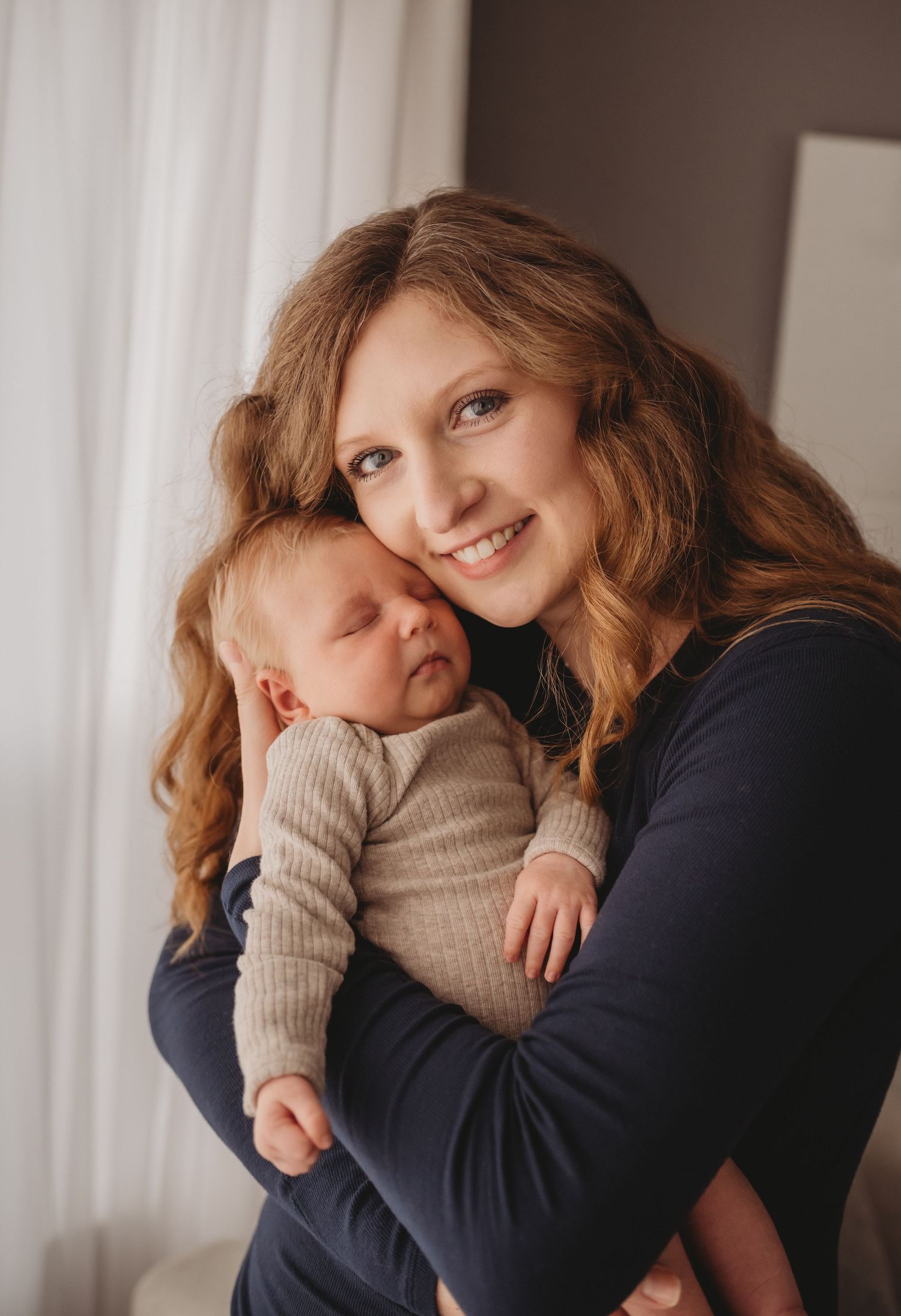 Smiling mother with curly hair holding sleeping newborn baby in cozy beige outfit indoors.