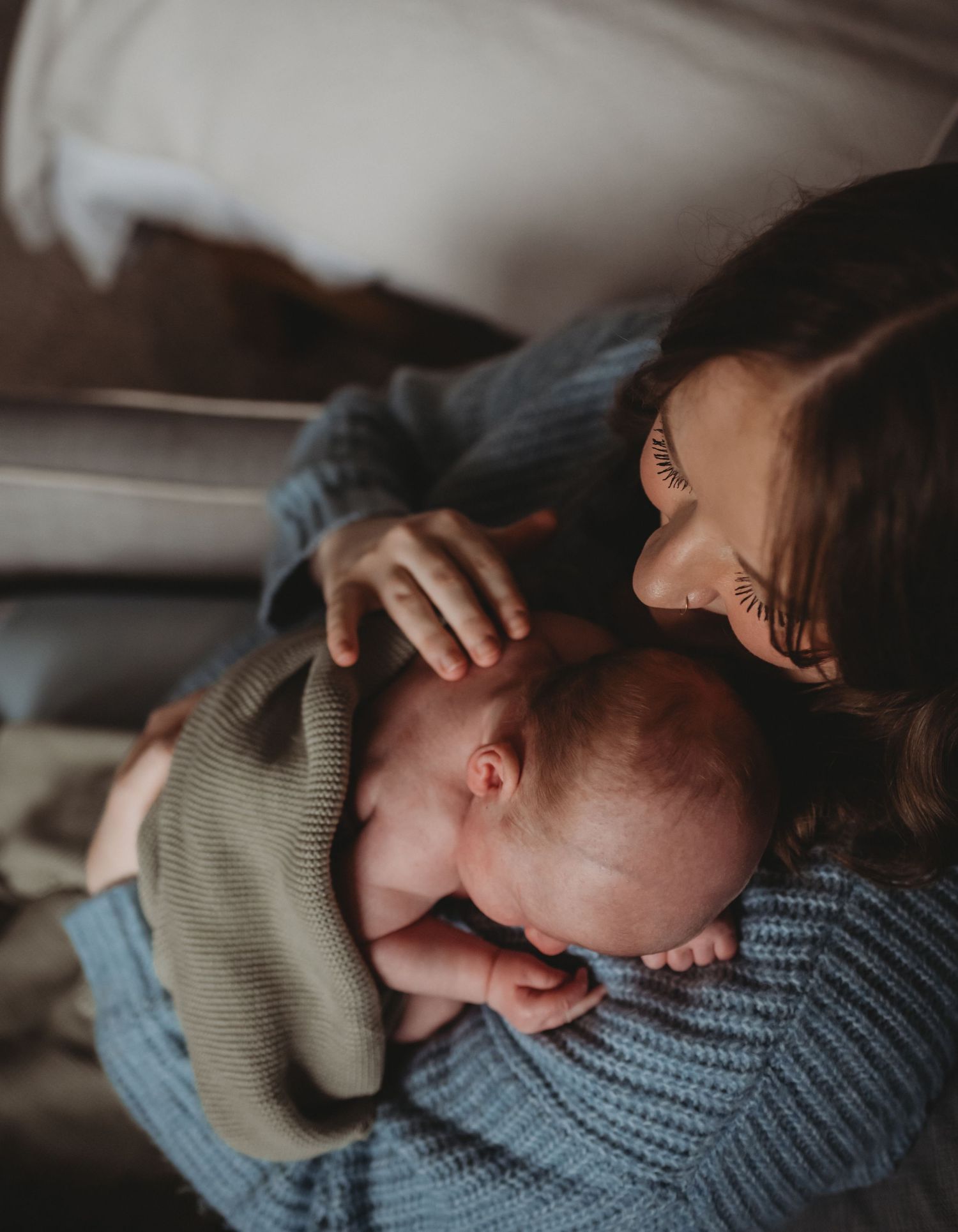 Mother in blue sweater gazing lovingly at her newborn baby cradled in her arms indoors.