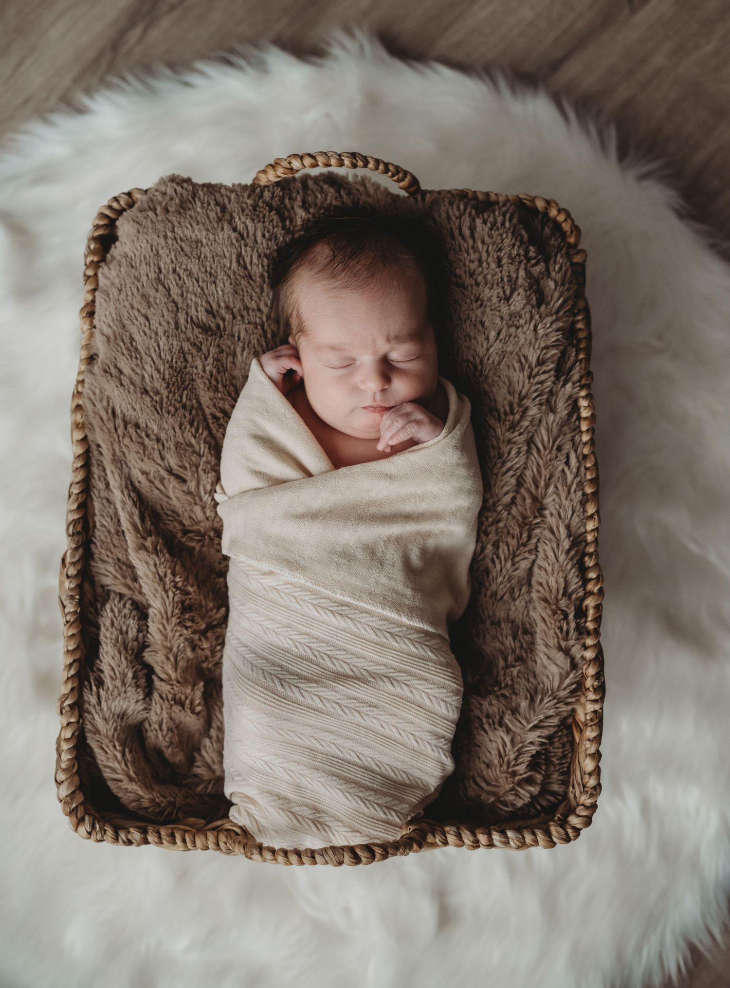 A swaddled newborn sleeps in a wicker basket on a white fluffy rug, viewed from above.