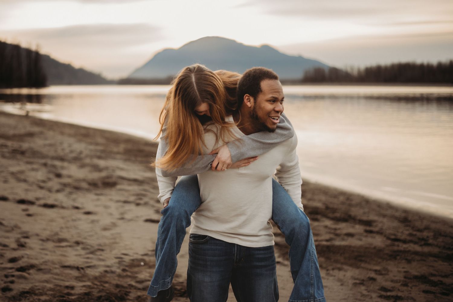 Happy interracial couple laughing as man gives woman a piggyback ride on a sandy lakeshore.