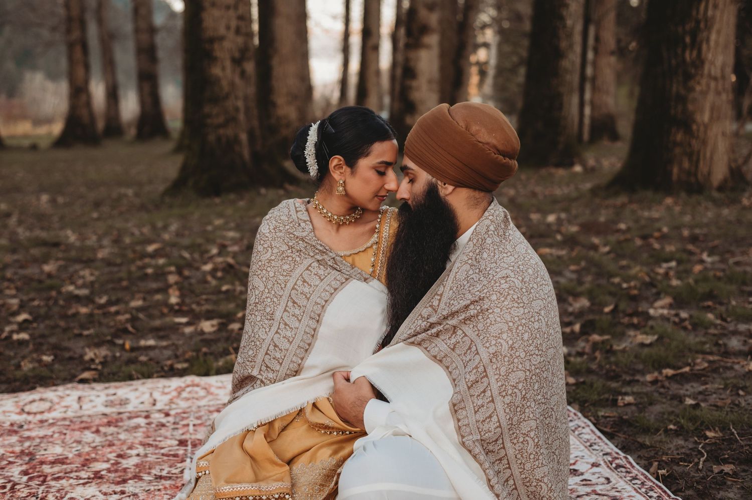 Sikh couple sharing an intimate moment wrapped together in a white shawl on a rug in a forest.