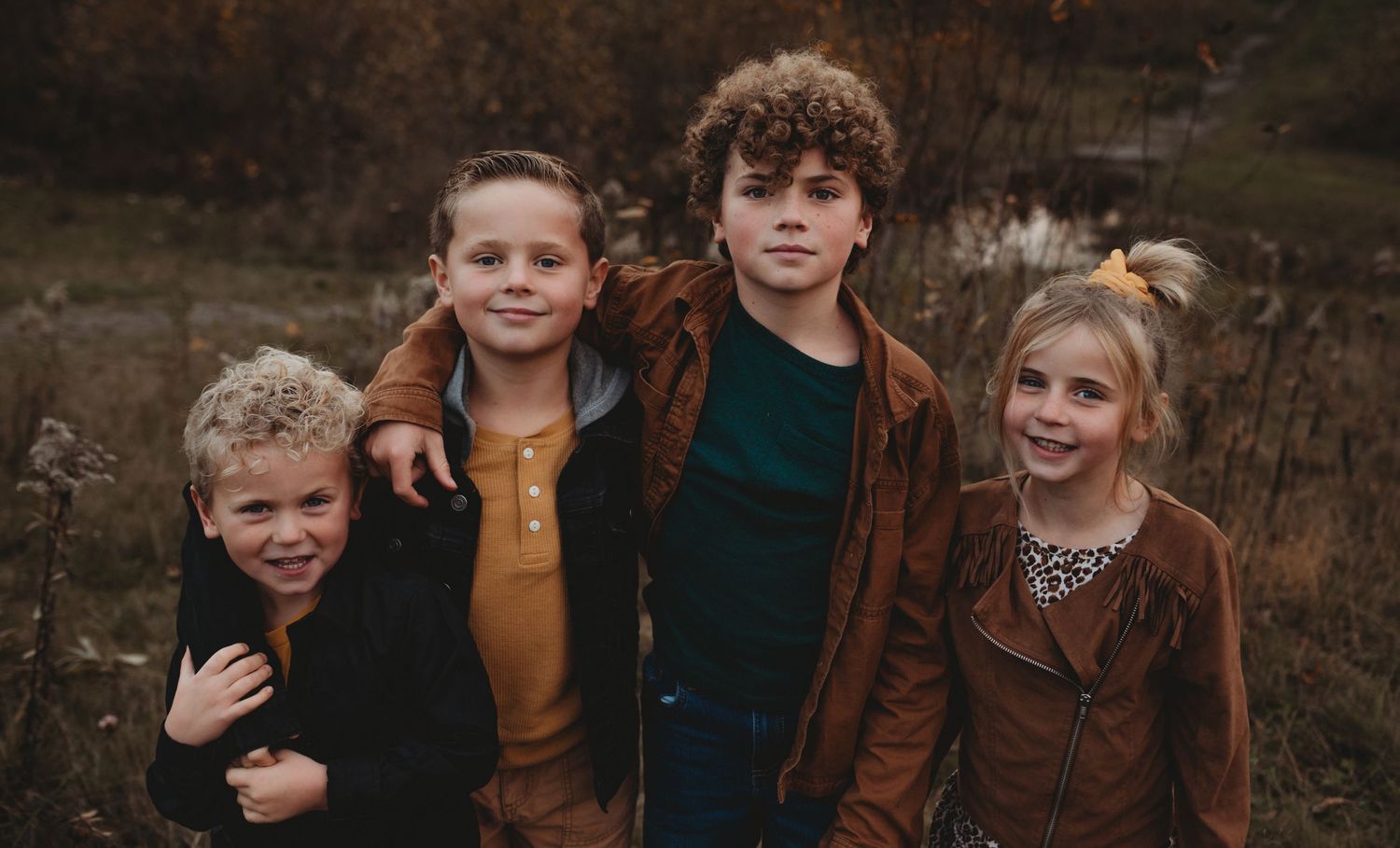 Four kids posing together in fall outfits in a rustic outdoor autumn setting.