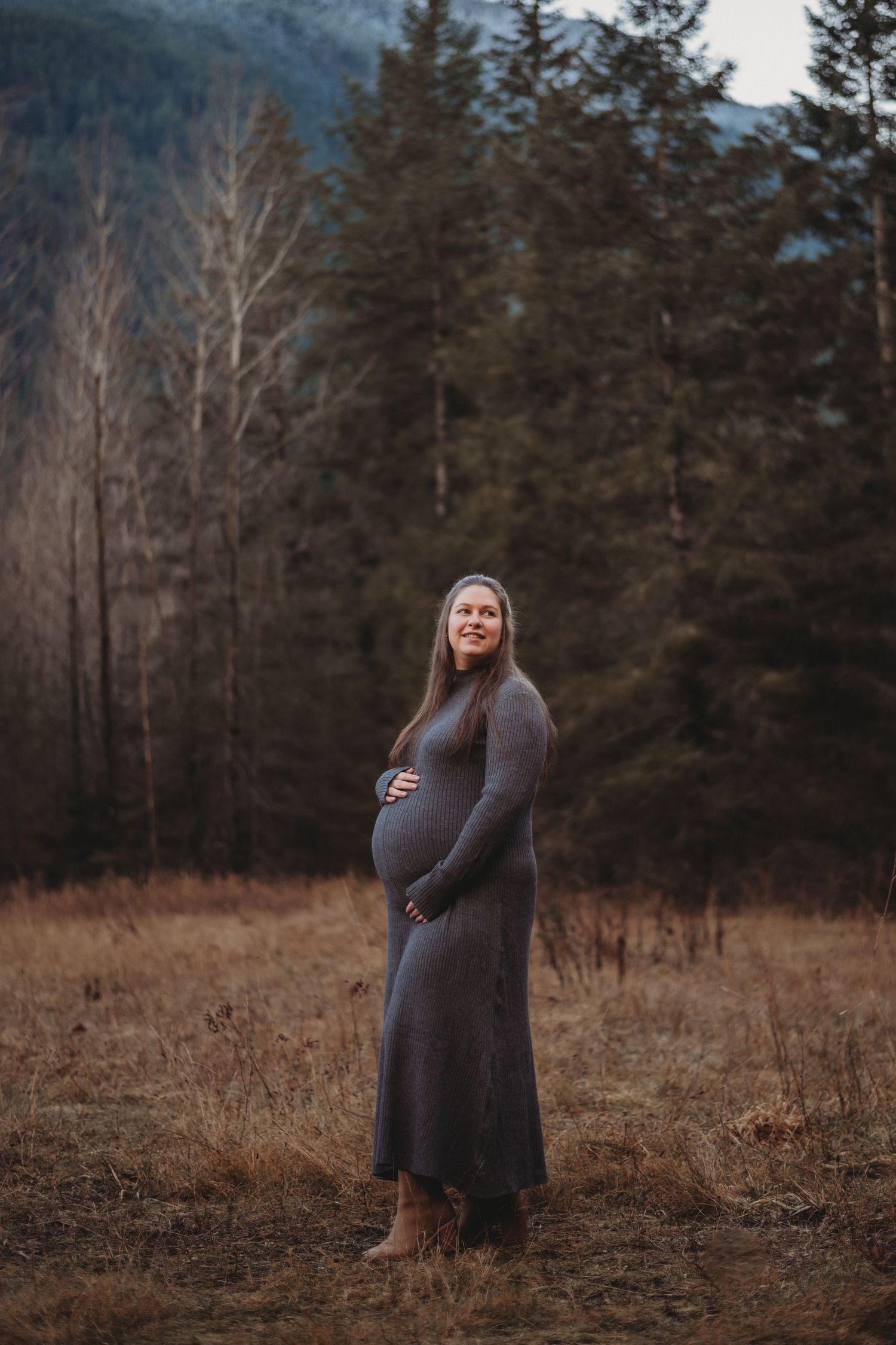 Pregnant woman in gray knit dress holding baby bump while standing in autumn forest meadow.