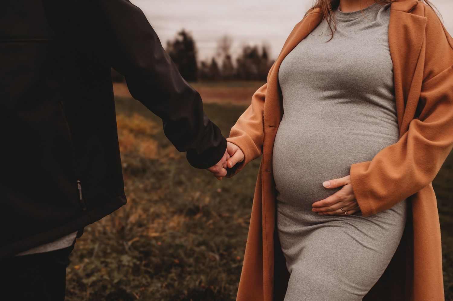Close-up of pregnant woman in gray dress and tan coat holding hands.